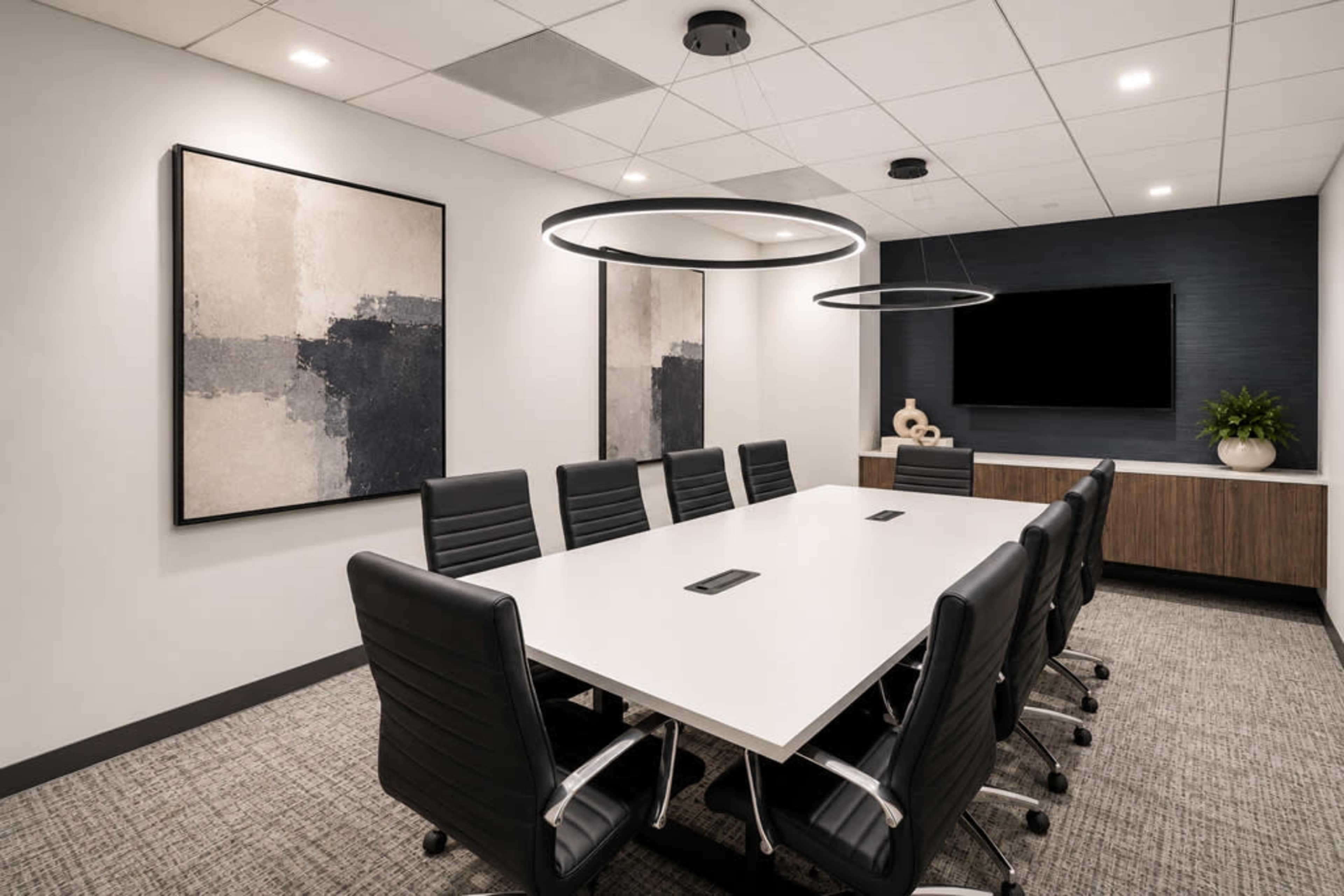 The image shows a modern conference room featuring a long white table surrounded by black office chairs, with abstract artwork on the walls and a television mounted on one side.