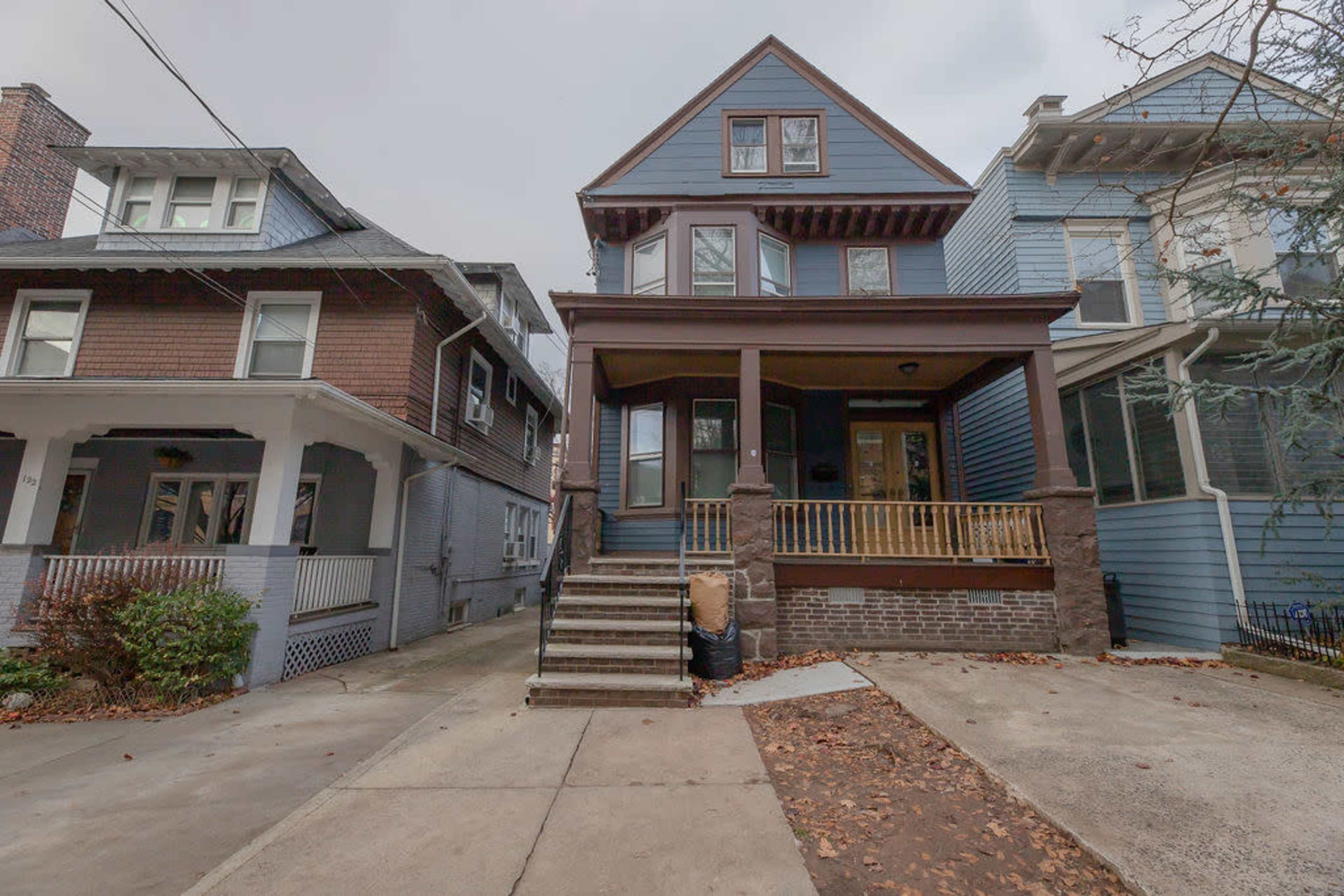 The image shows a blue two-and-a-half-story house with a front porch, flanked by two neighboring homes, along a concrete driveway.