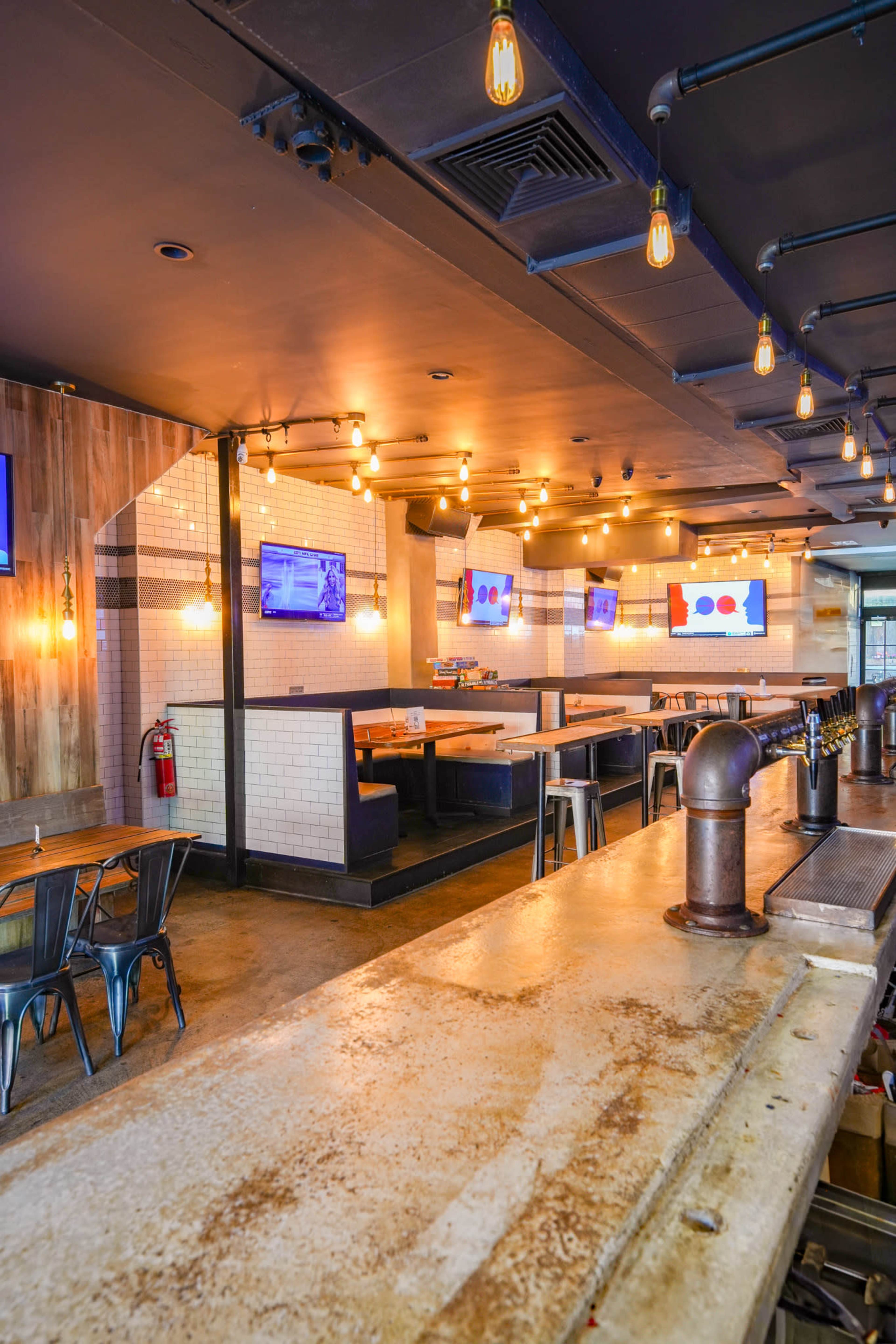 The interior of a modern restaurant features industrial-style decor with wooden accents, hanging light bulbs, and several televisions mounted on the walls.