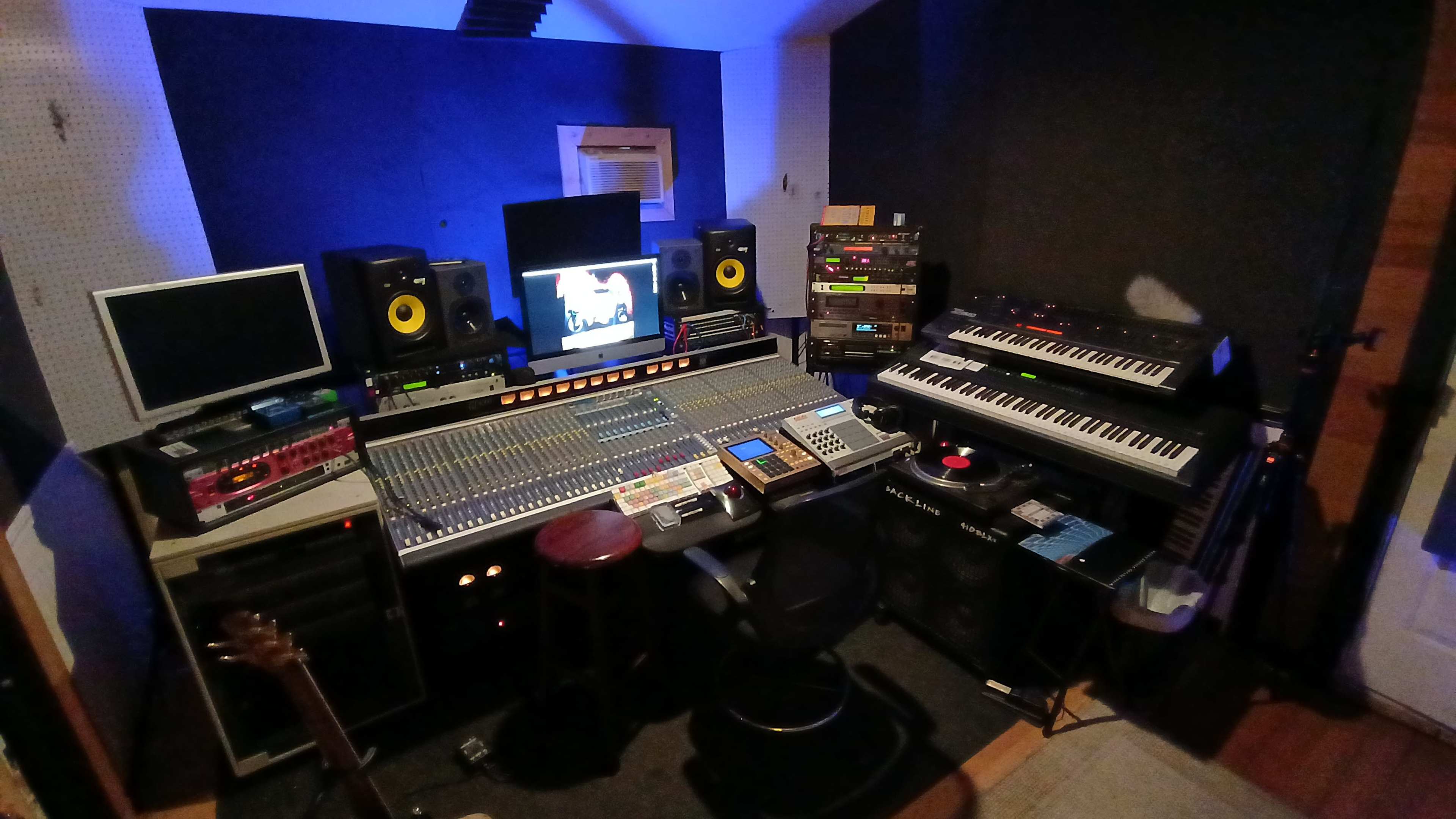 The image shows a recording studio with a large mixing console, multiple keyboards, computer monitors, and various audio equipment arranged around the room.