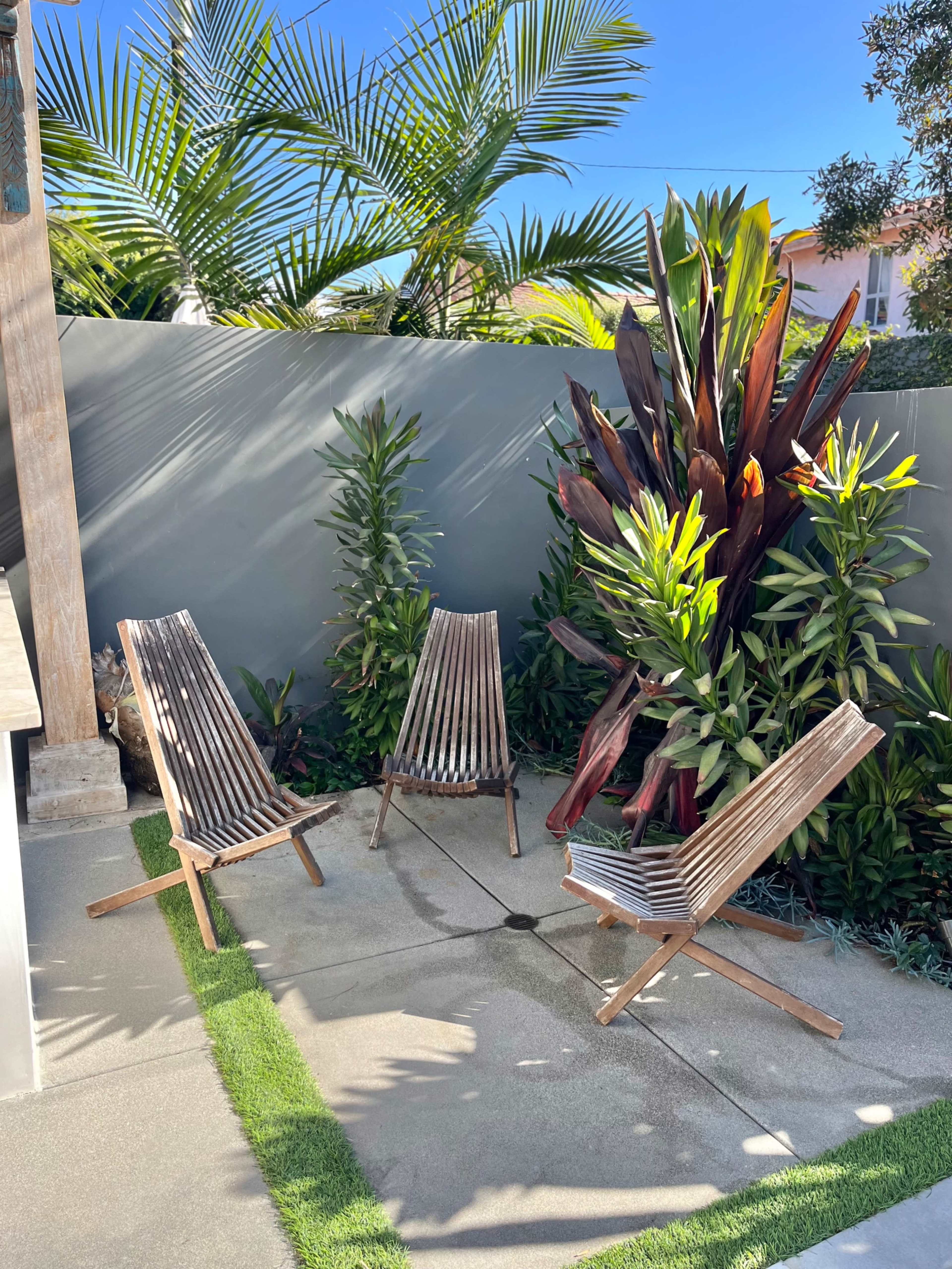 Three wooden lounge chairs are positioned on a concrete patio surrounded by lush tropical plants and palm trees.