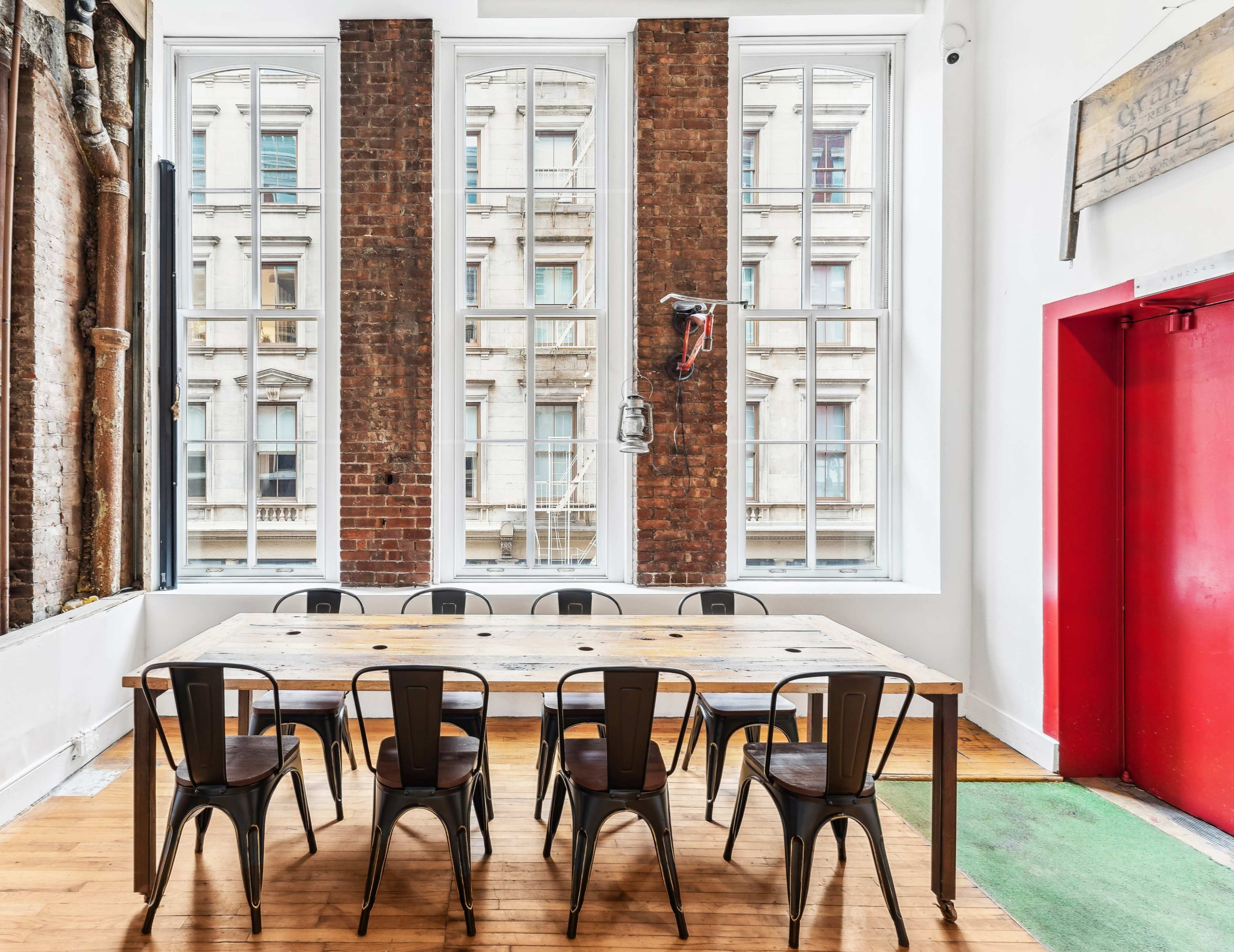 A long wooden table surrounded by metal chairs is positioned in a brightly lit room with large windows and exposed brick walls.