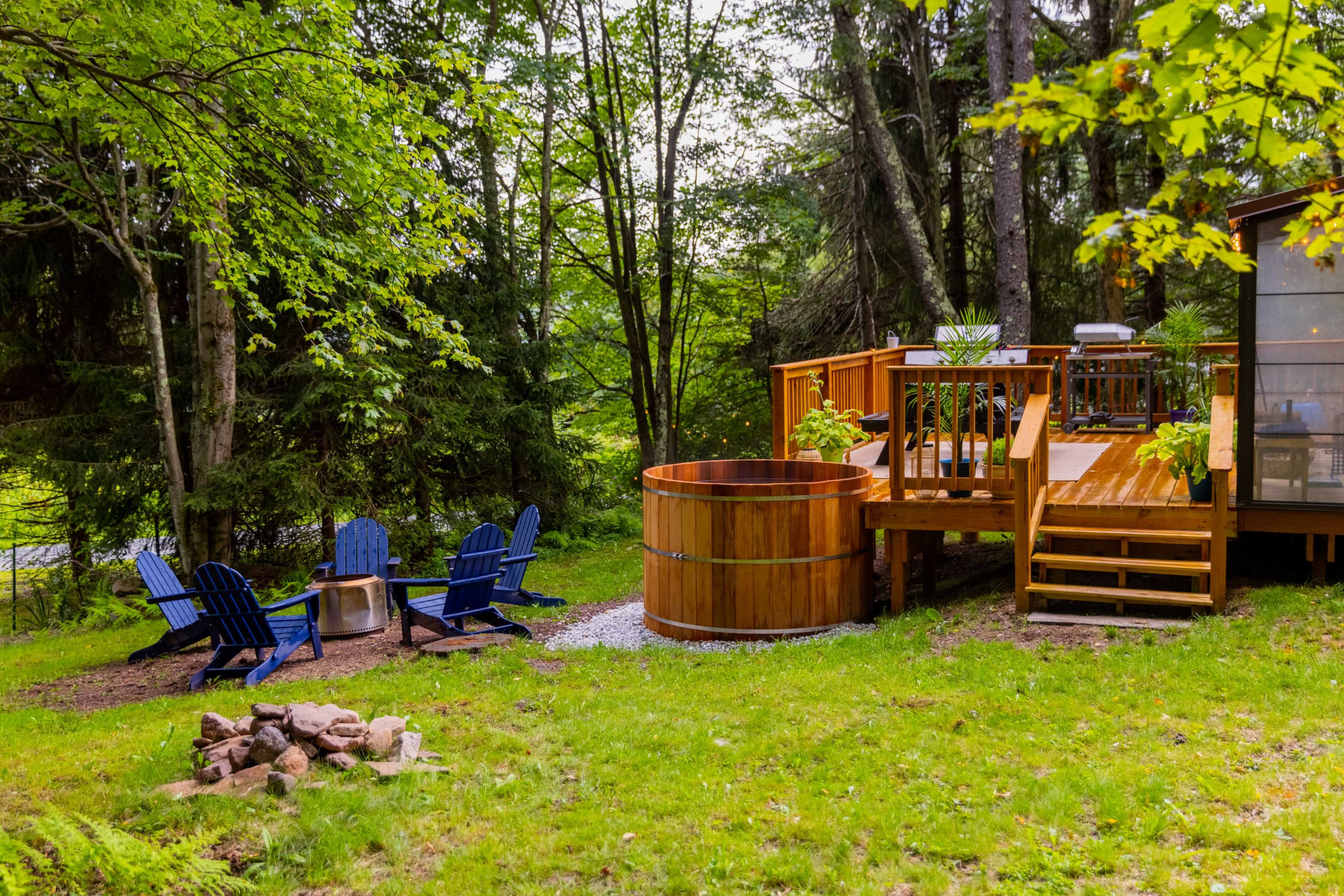 A wooden deck with a hot tub and seating area surrounded by trees in a grassy outdoor setting.
