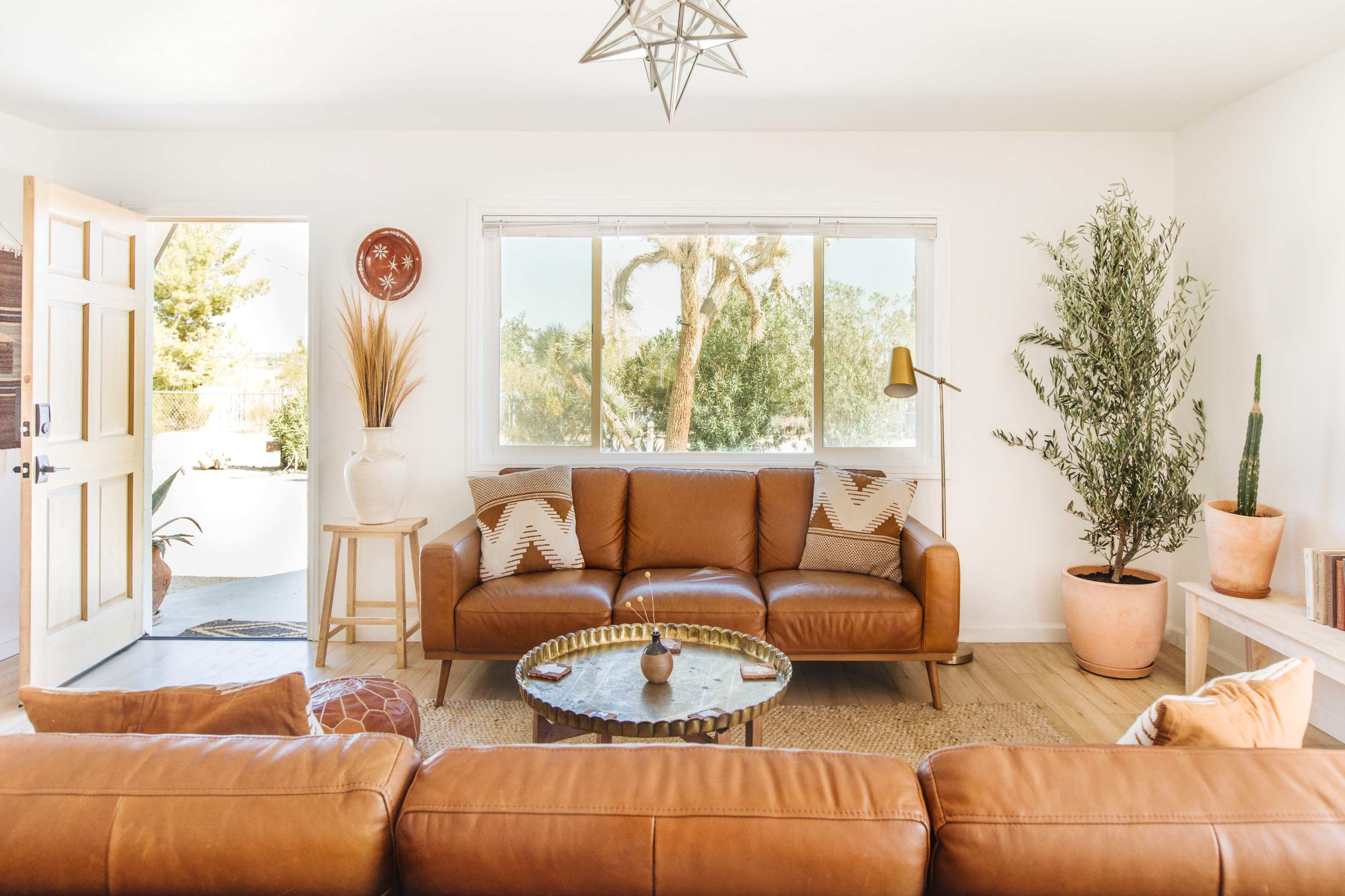 A modern living room features a brown leather sofa, a round coffee table, and decorative plants, with a door leading to an outdoor space.