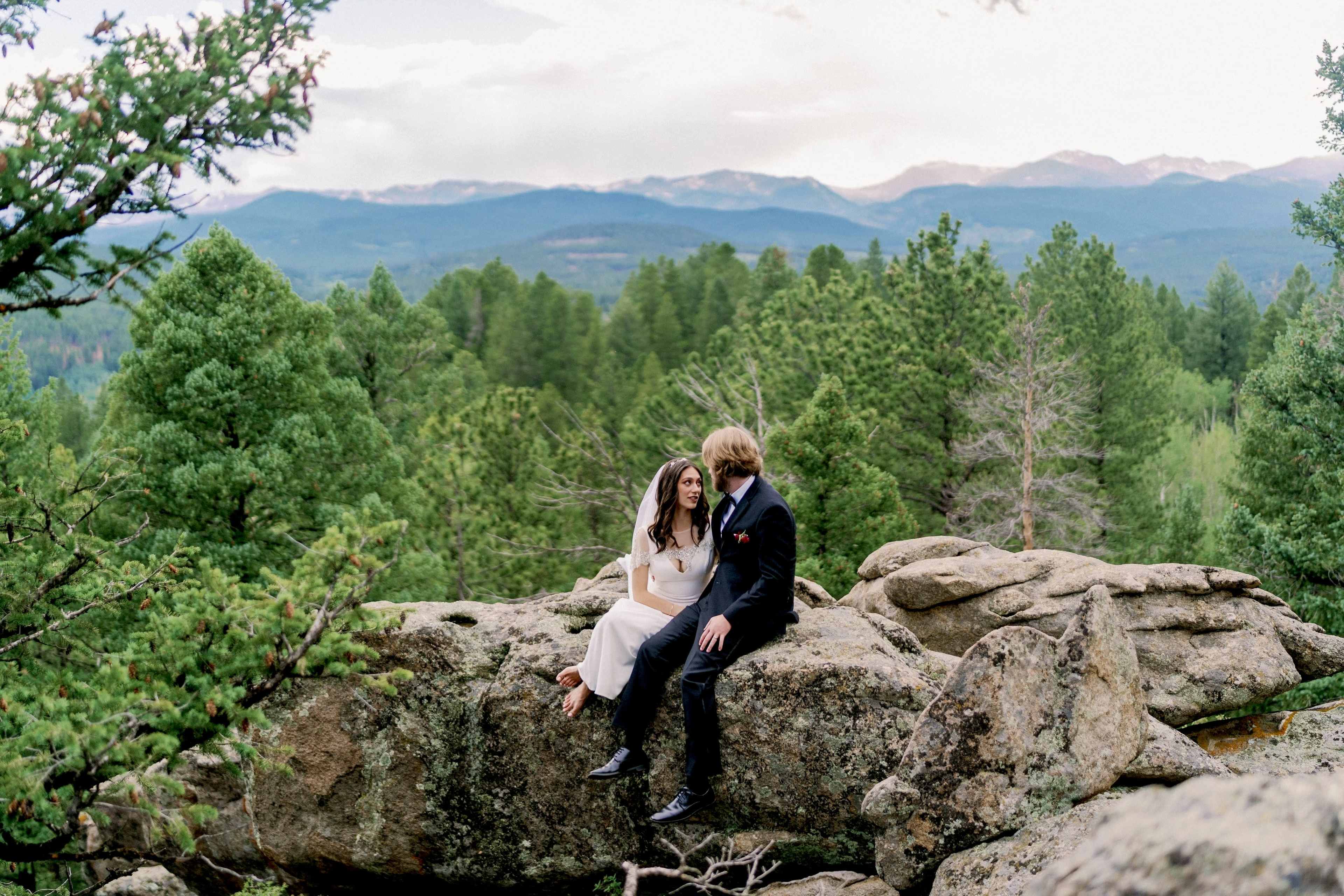 A couple sits together on a large rock surrounded by pine trees and mountainous terrain.