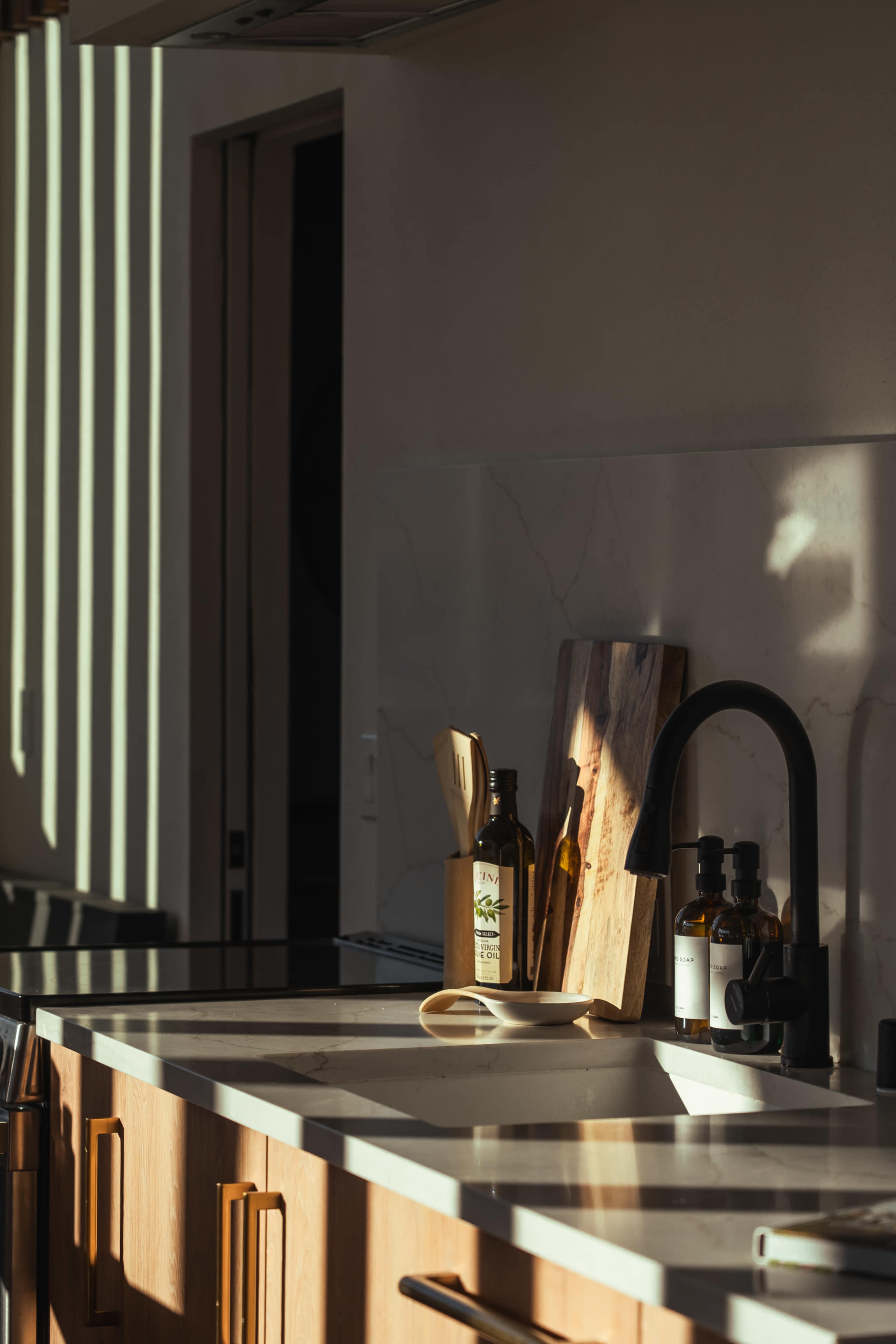 A kitchen countertop is bathed in natural light with shadows cast by vertical blinds, featuring olive oil bottles and a wooden cutting board.