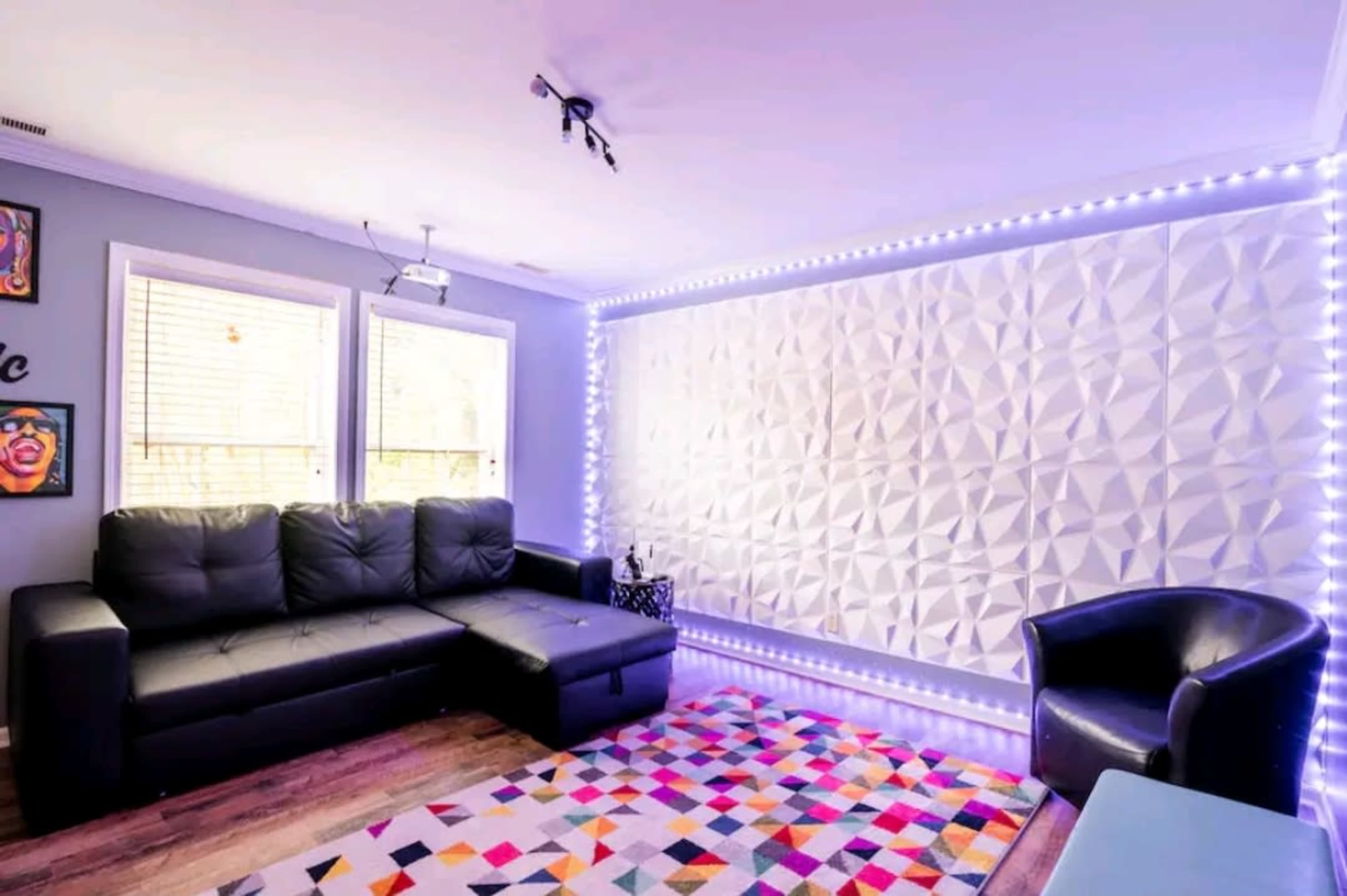 A modern living room features a sectional black sofa, a black armchair, a patterned area rug, and a textured wall illuminated with LED lights.