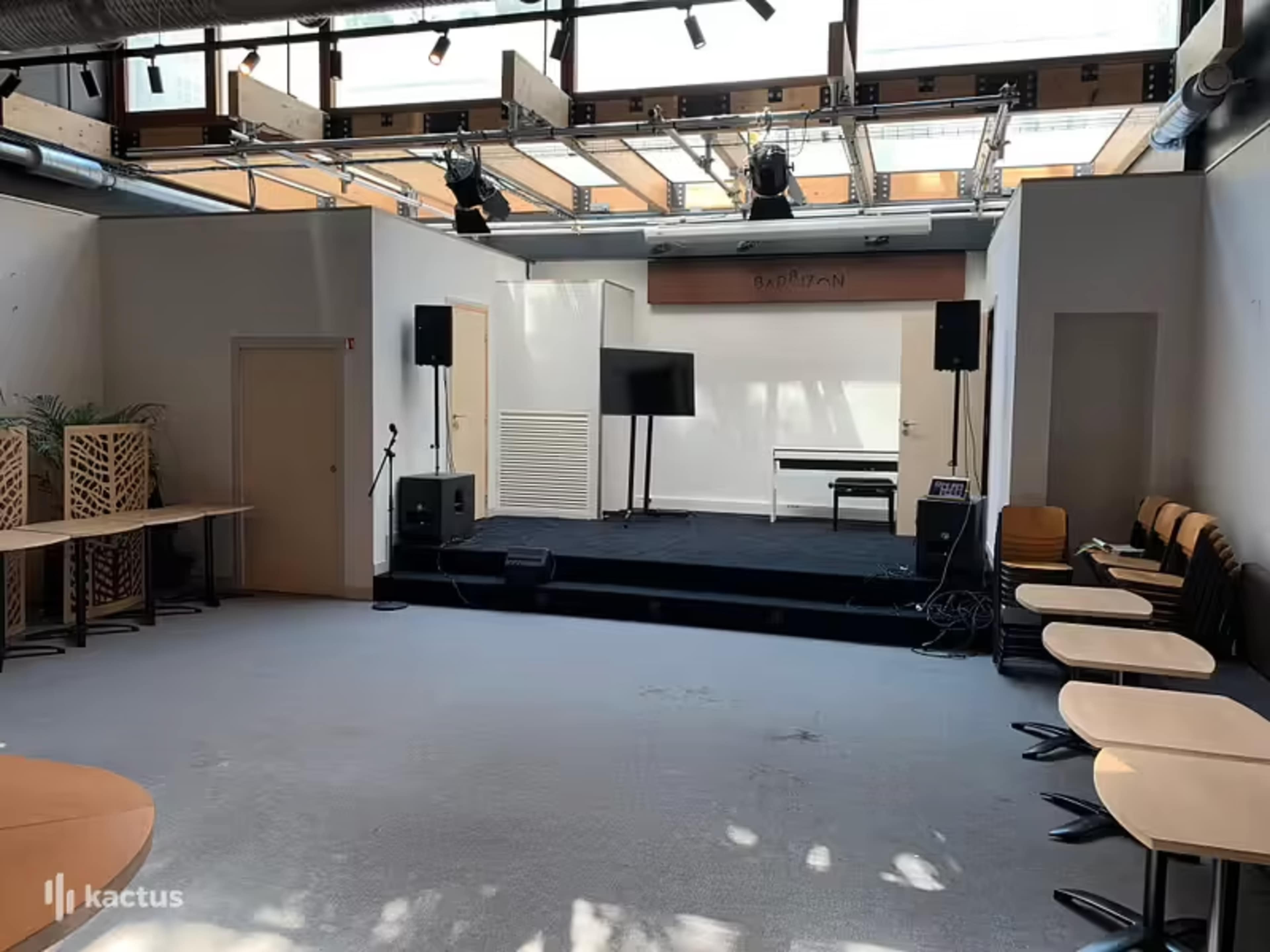 An empty indoor event space featuring a stage with a microphone, two speakers, a television screen, and rows of chairs arranged in a semicircle.