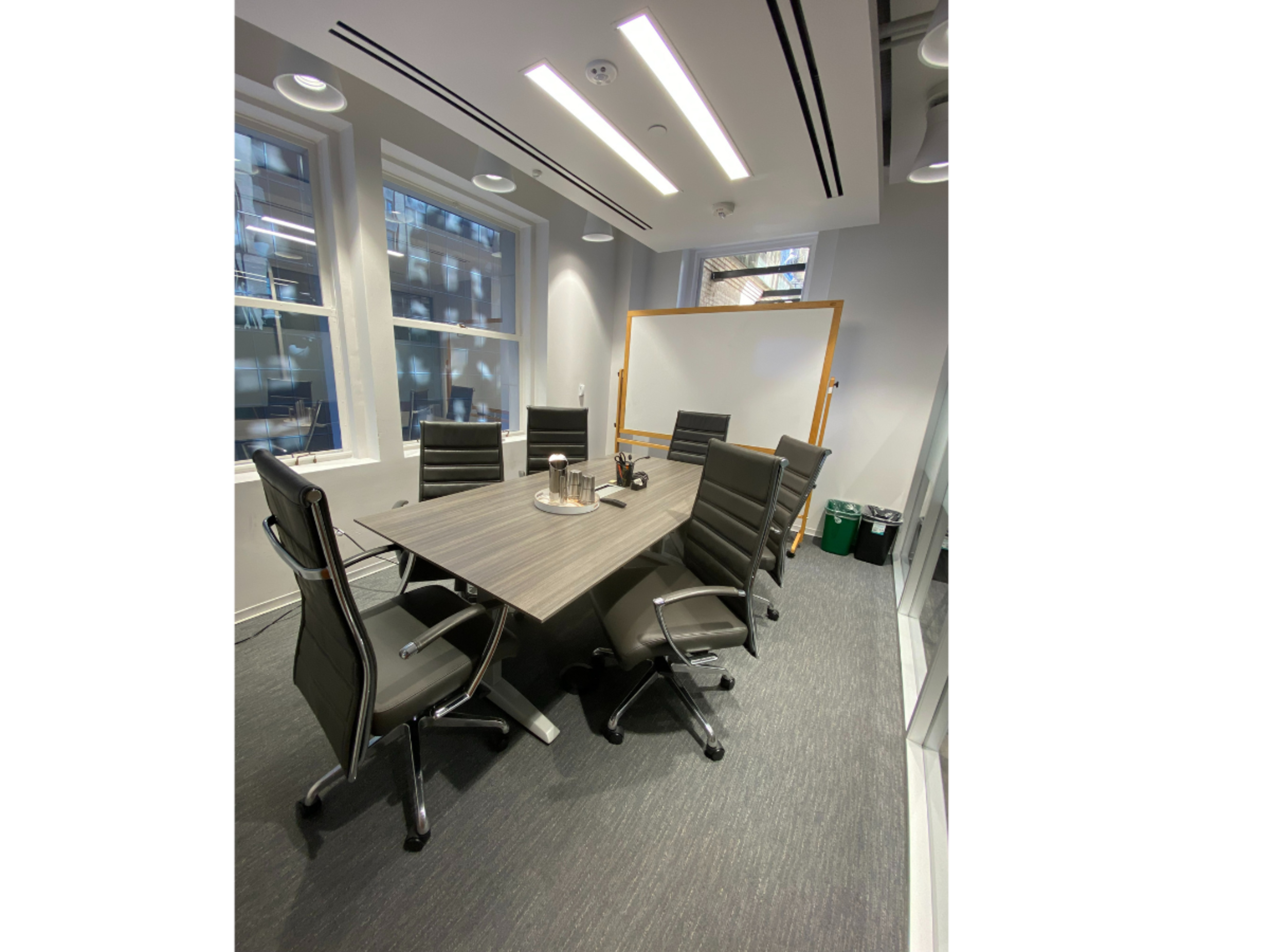 A modern conference room features a long table surrounded by ergonomic chairs, with large windows and a whiteboard in the background.