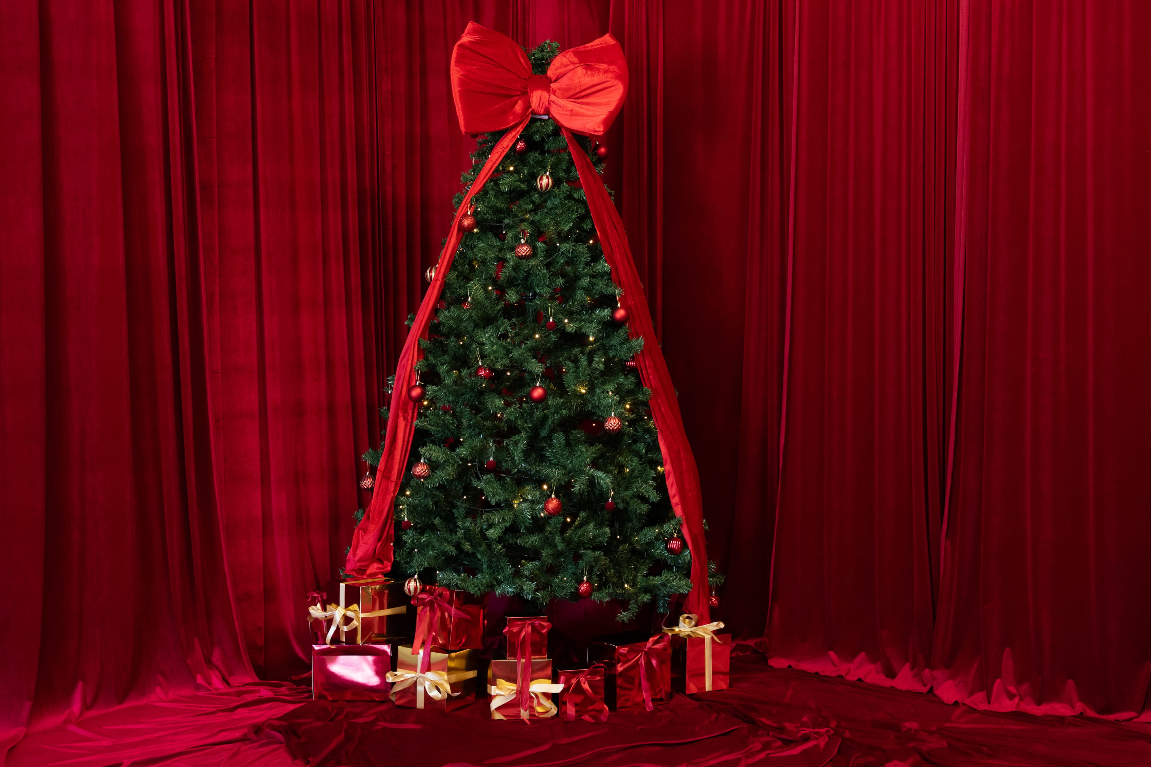 A decorated Christmas tree with a large red bow stands against a backdrop of red curtains, surrounded by neatly wrapped presents.