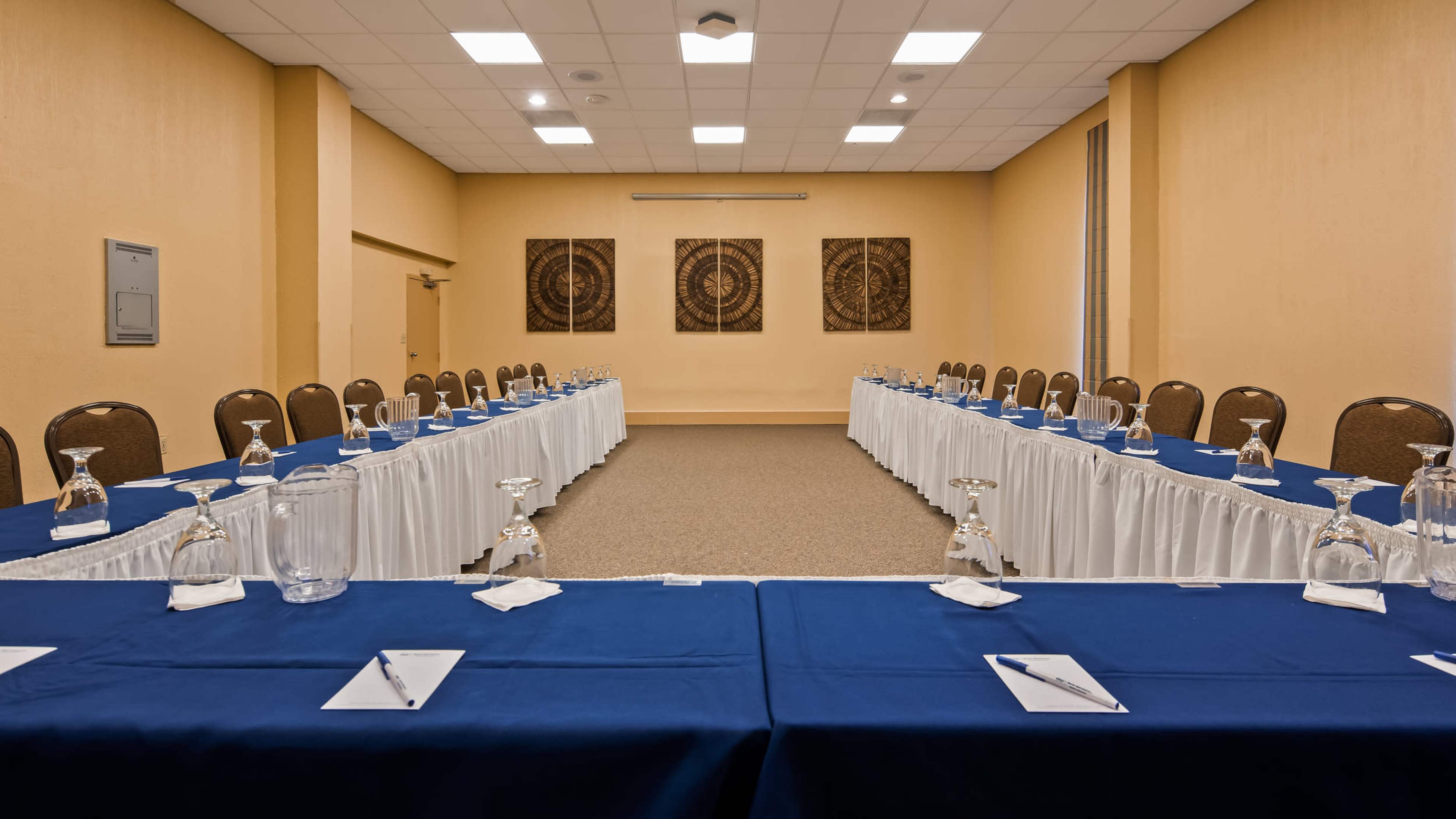 A conference room set up with two long tables covered in blue tablecloths, facing each other and bordered by chairs, with decorative wall art in the background.