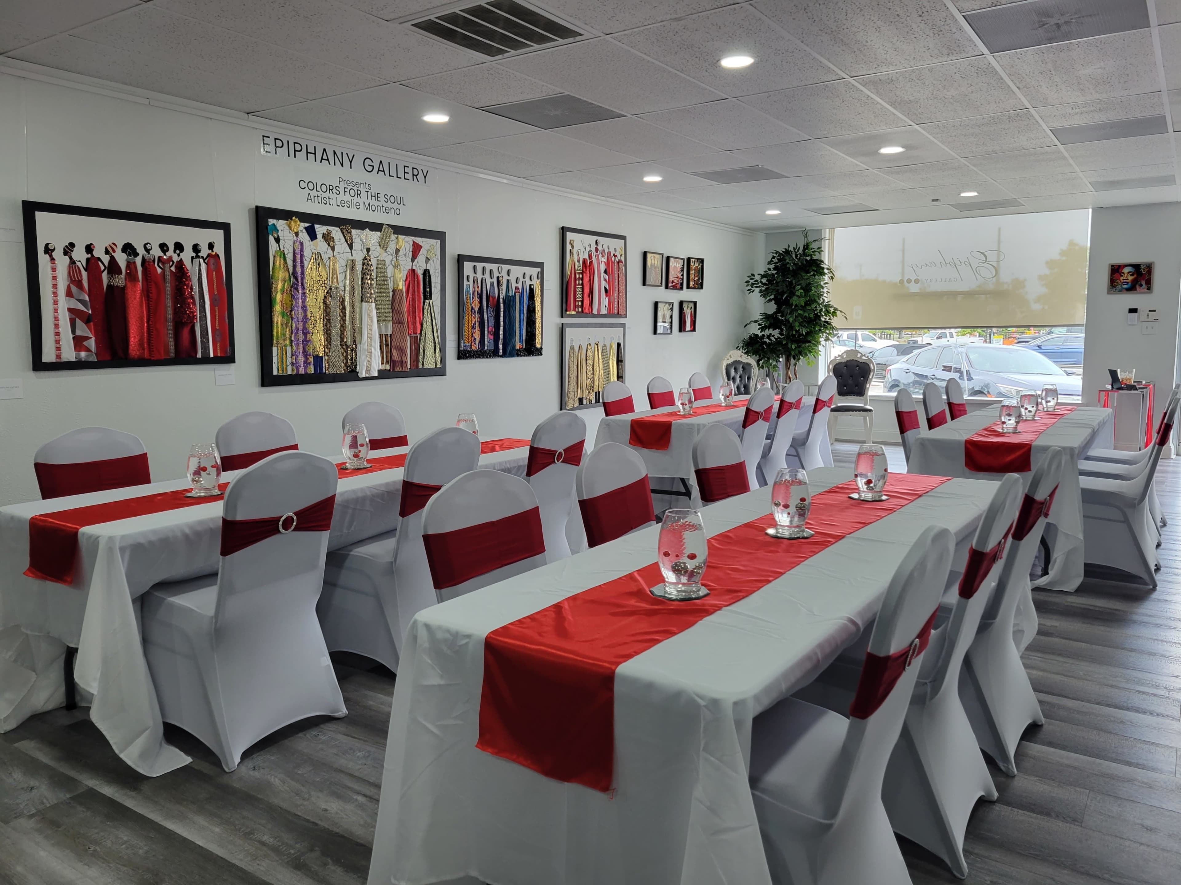 A well-decorated event space with tables covered in white cloths and red runners, surrounded by framed artwork on the walls.