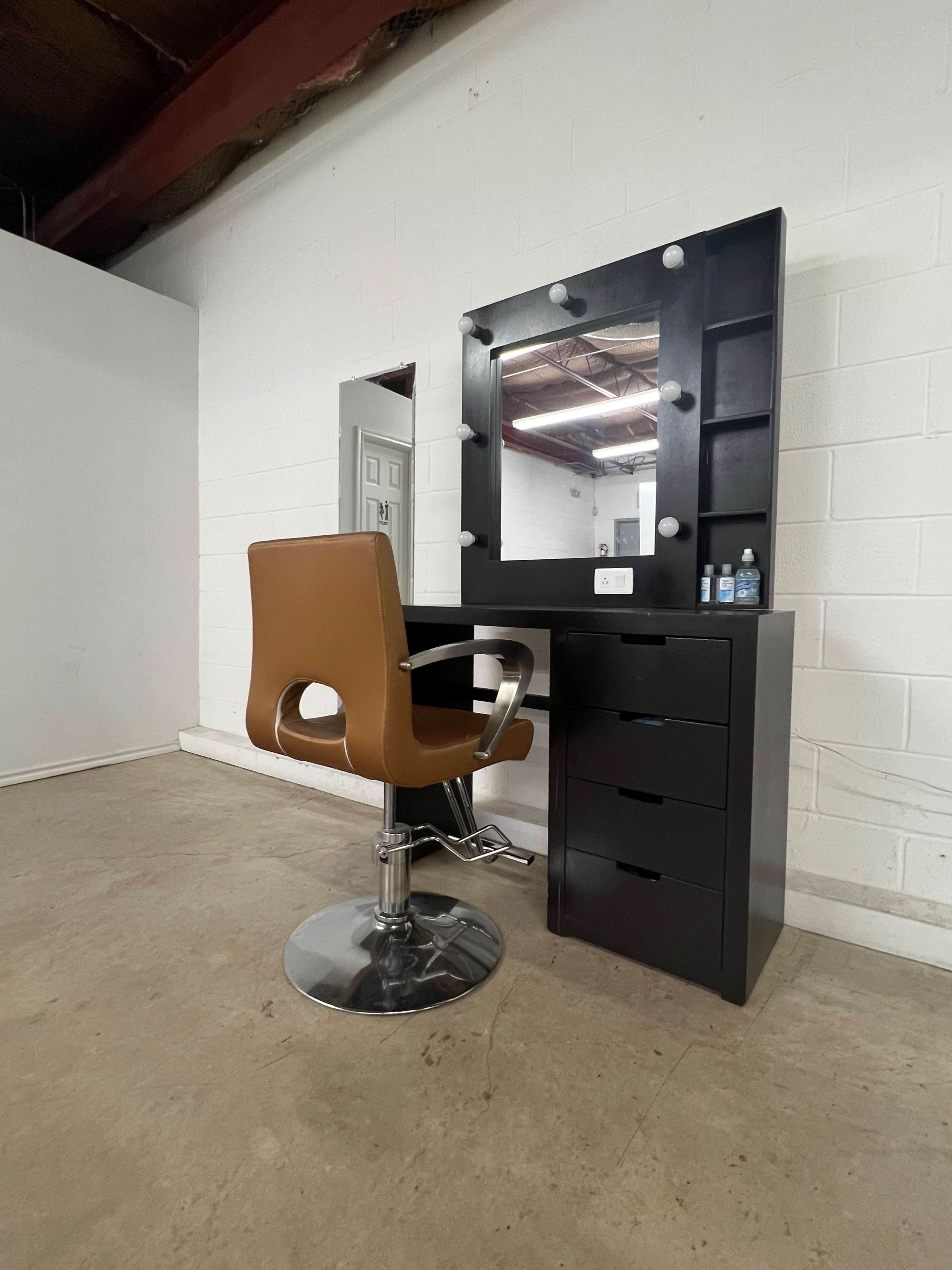 A modern makeup vanity with a large mirror and a swivel chair, positioned against a gray wall in a sparse room.