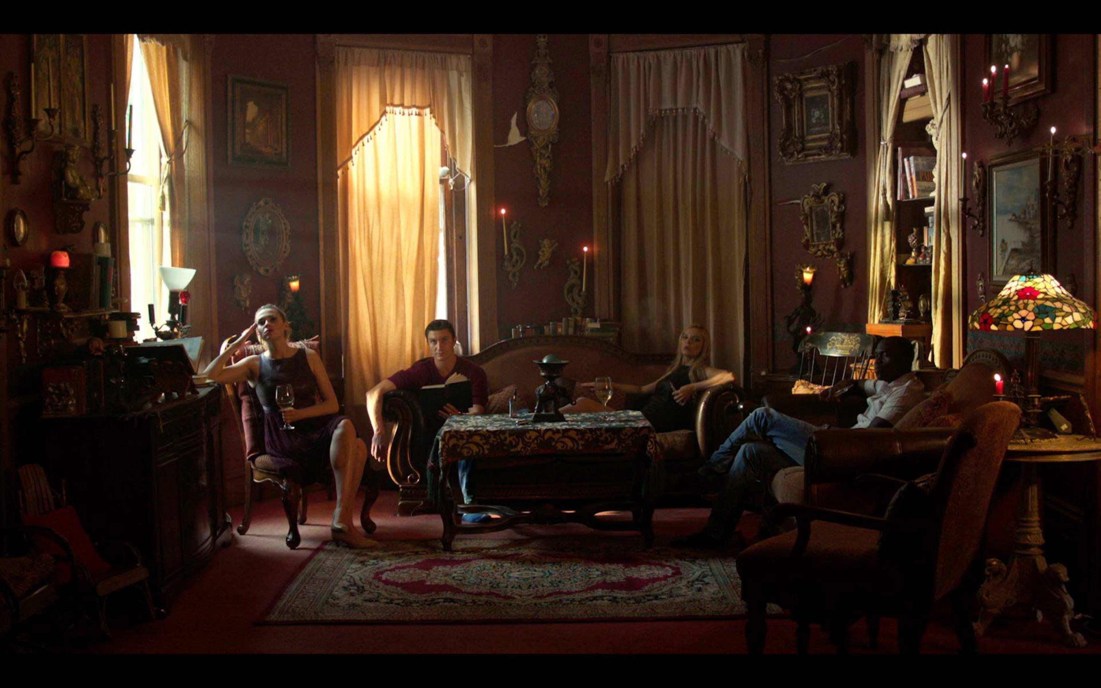 A group of four people sits in a dimly lit, ornately decorated room with vintage furnishings and large windows.