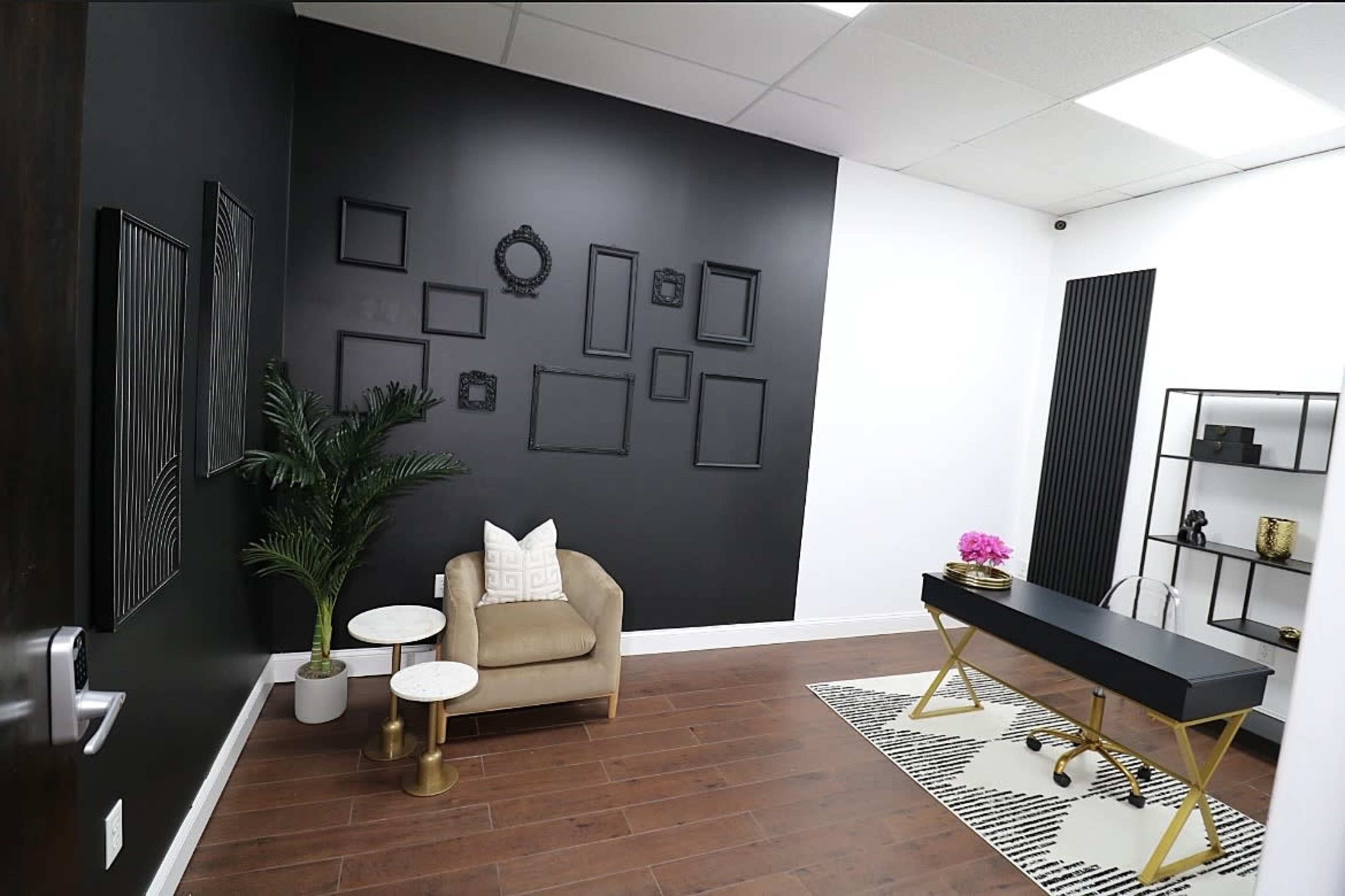 A modern office space featuring a black accent wall with empty picture frames, a beige armchair next to two small tables, and a black desk positioned on a patterned rug.