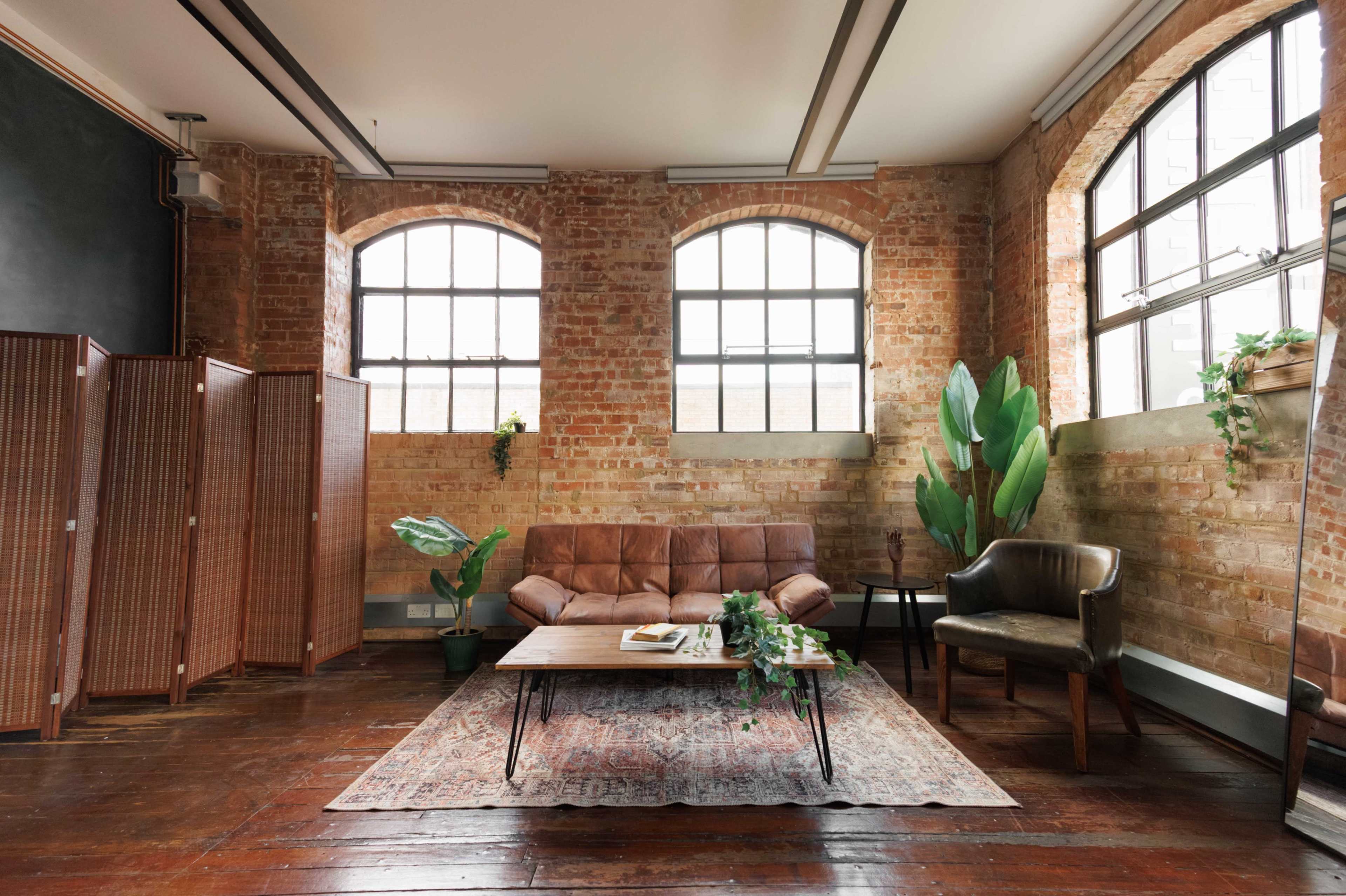 The image shows a stylish living room with exposed brick walls, a brown leather sofa, a wooden coffee table, and large windows allowing natural light.
