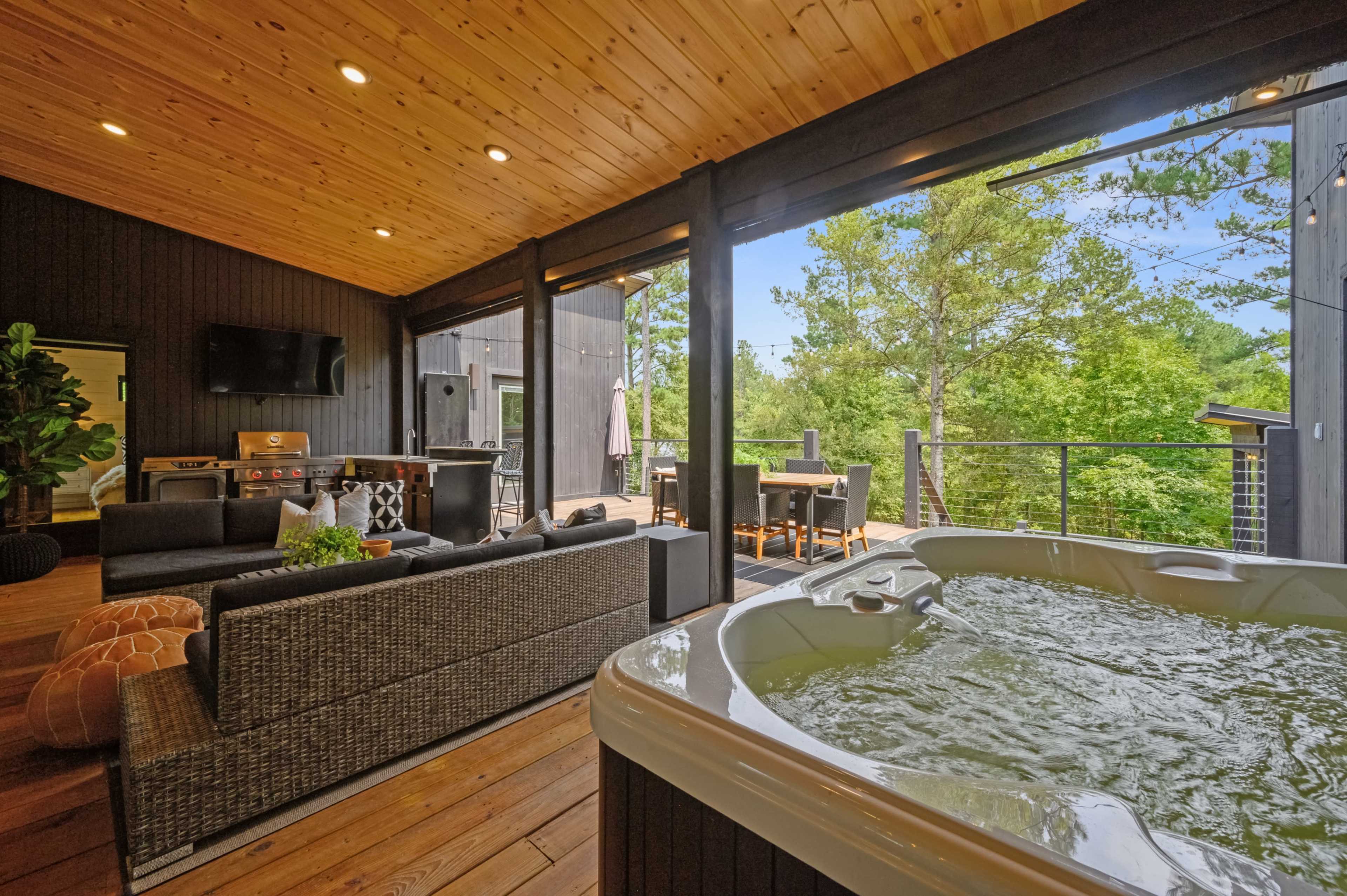 A lounge area featuring a hot tub, comfortable seating, and a dining table on a covered porch overlooking greenery.