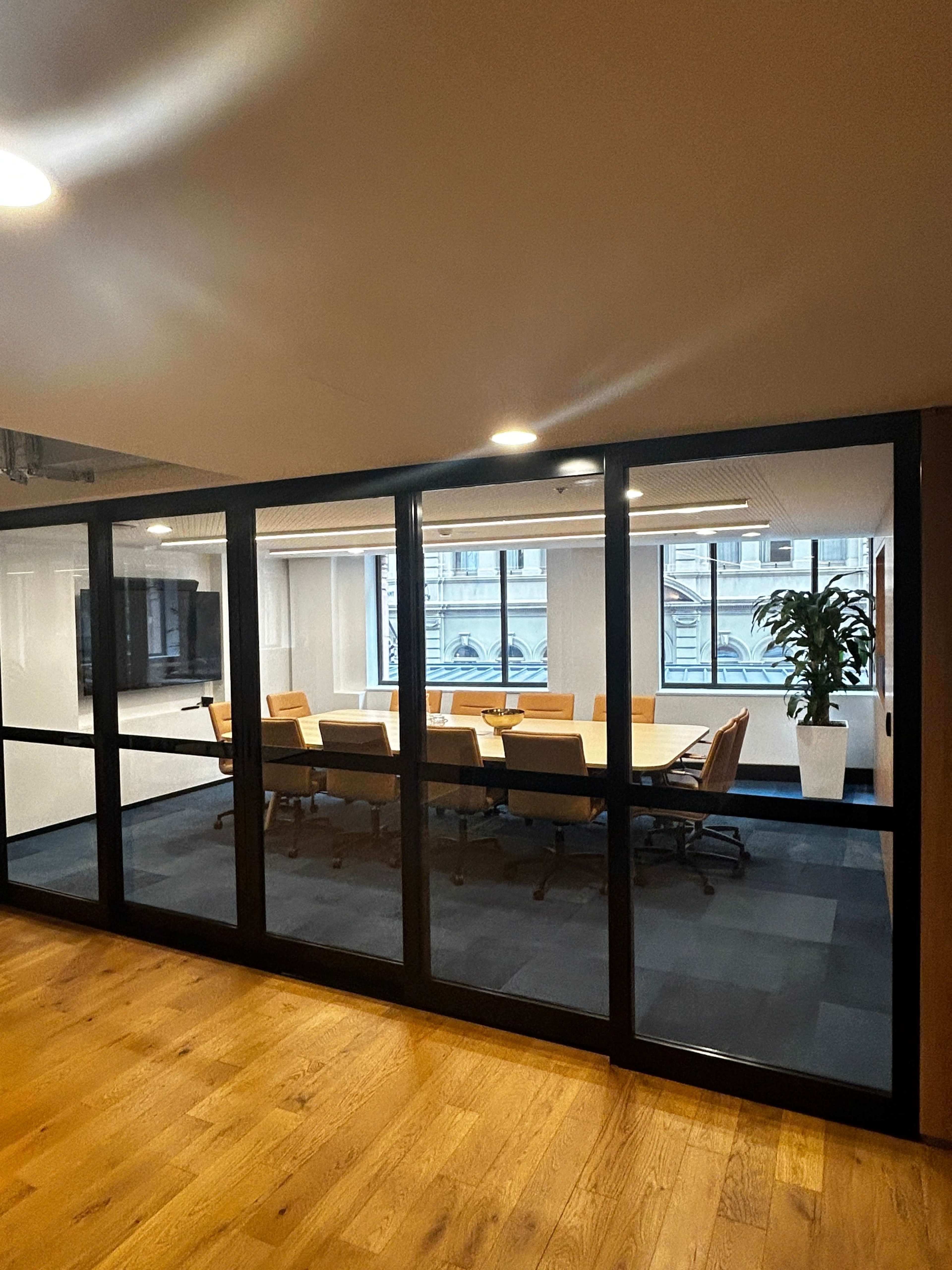 A modern conference room with a large table and multiple chairs is visible through glass walls, featuring a plant and a television on the wall.