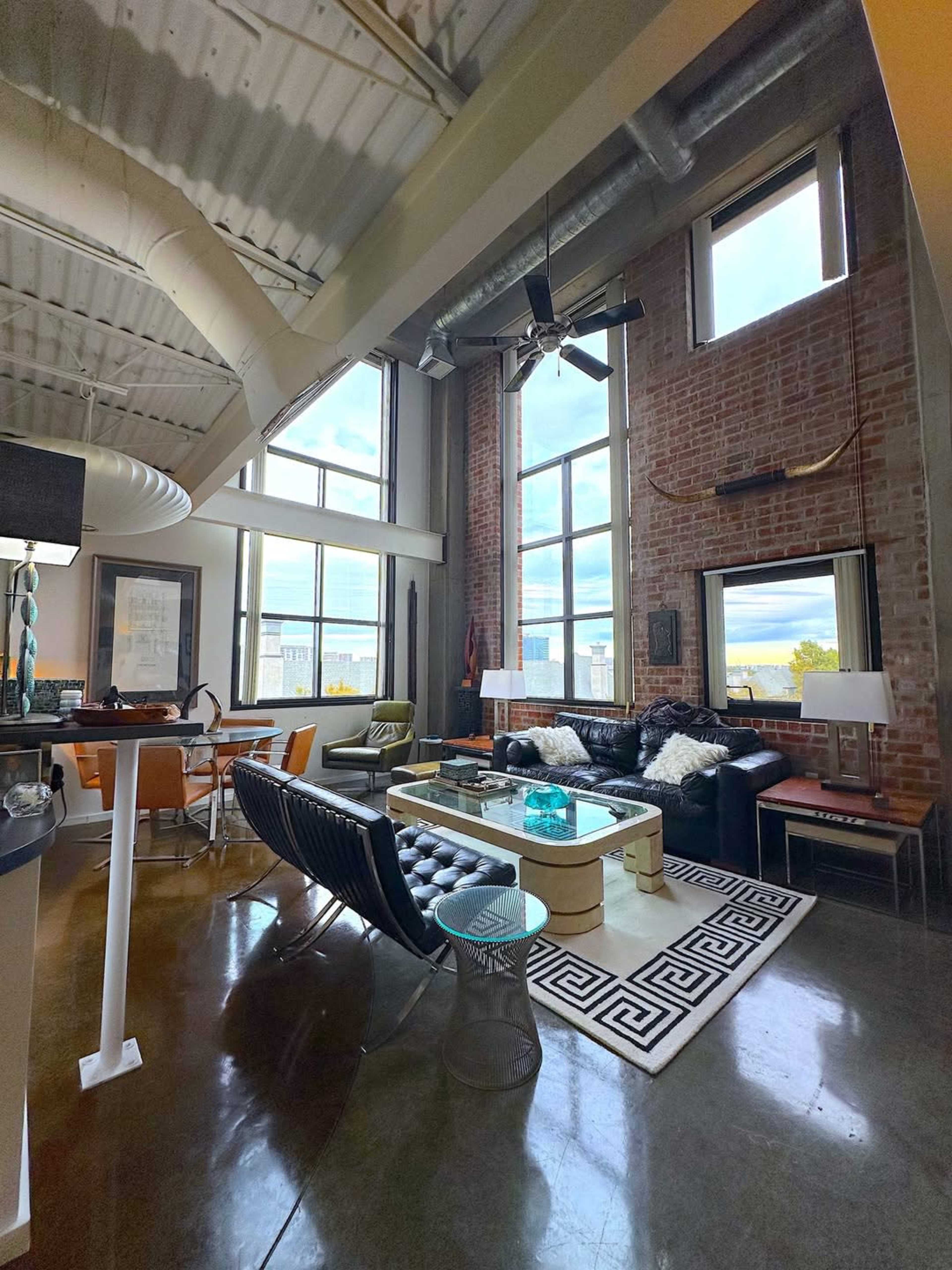 The image shows a modern loft-style living room with large windows, exposed brick walls, and industrial decor.