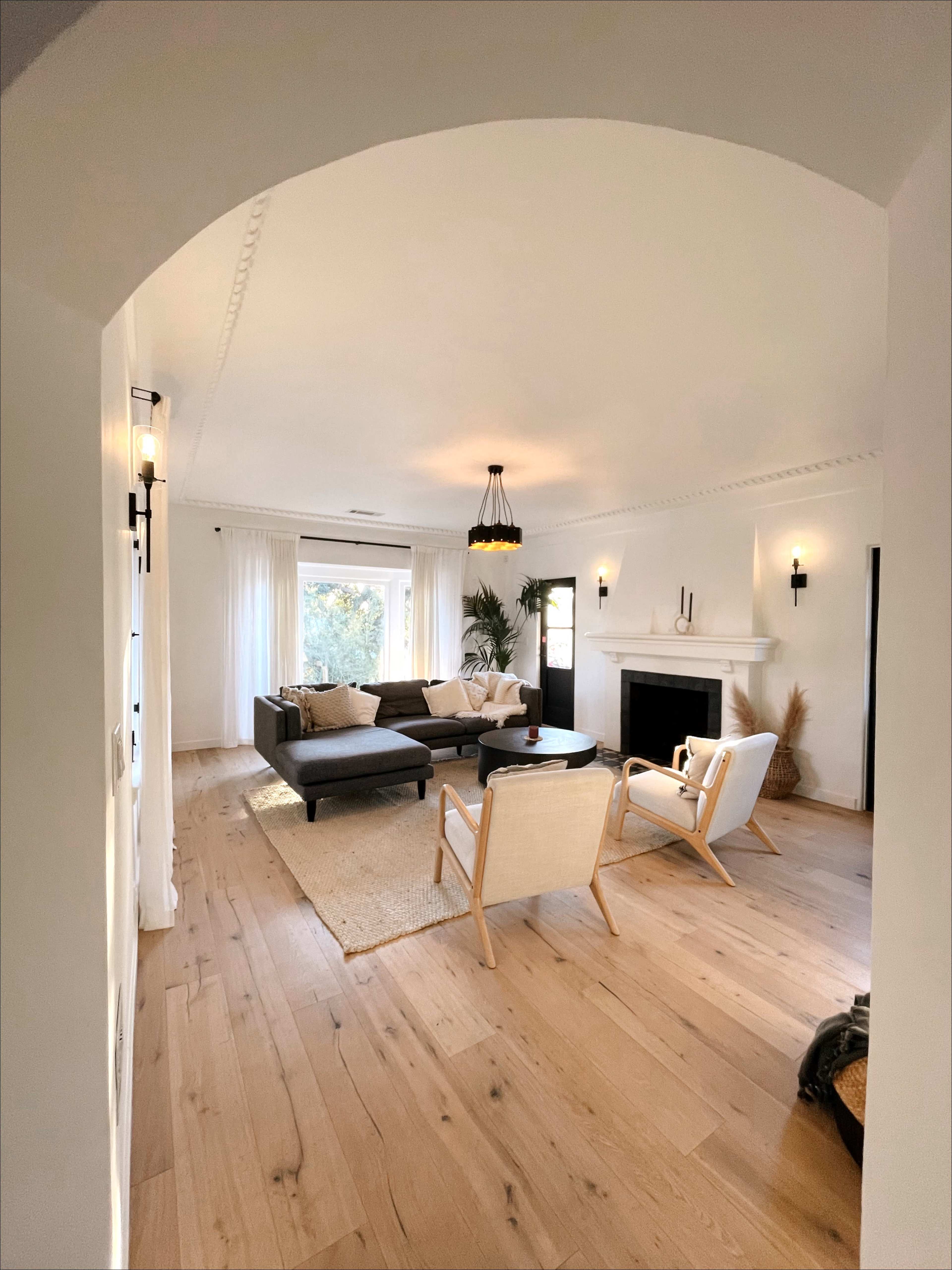 The image shows a light and airy living room featuring a black sectional sofa, two white chairs, a fireplace, and large windows with white curtains.