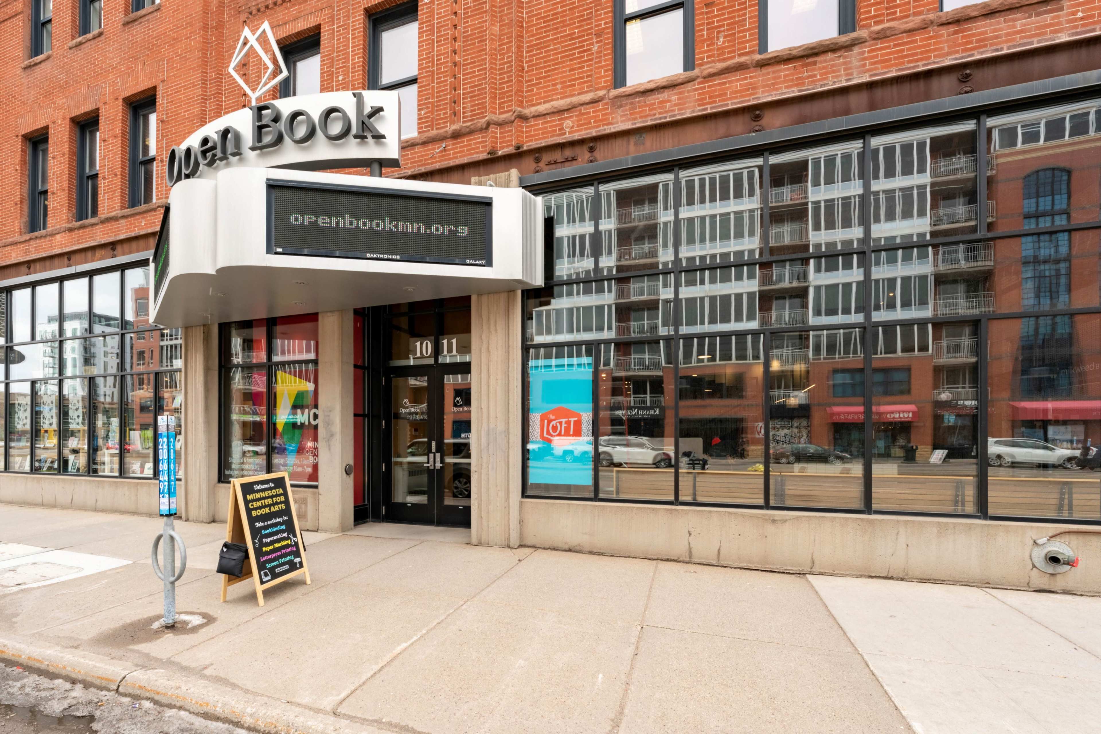 The entrance of the Open Book building features a sign with its website and a chalkboard on the sidewalk, set against a backdrop of brick architecture and large windows.