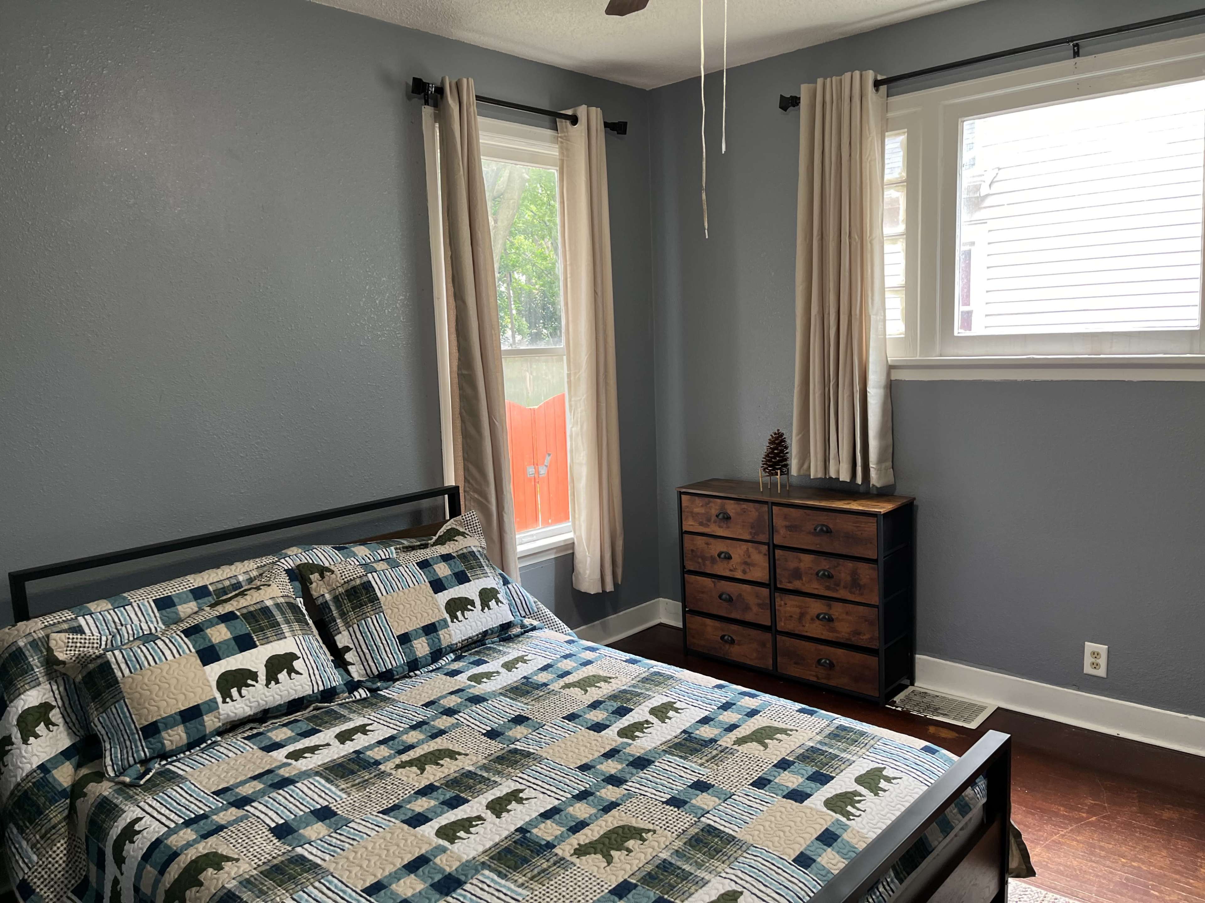 A bedroom features a metal frame bed with a patterned quilt, a wooden dresser, and two windows with light-colored curtains.