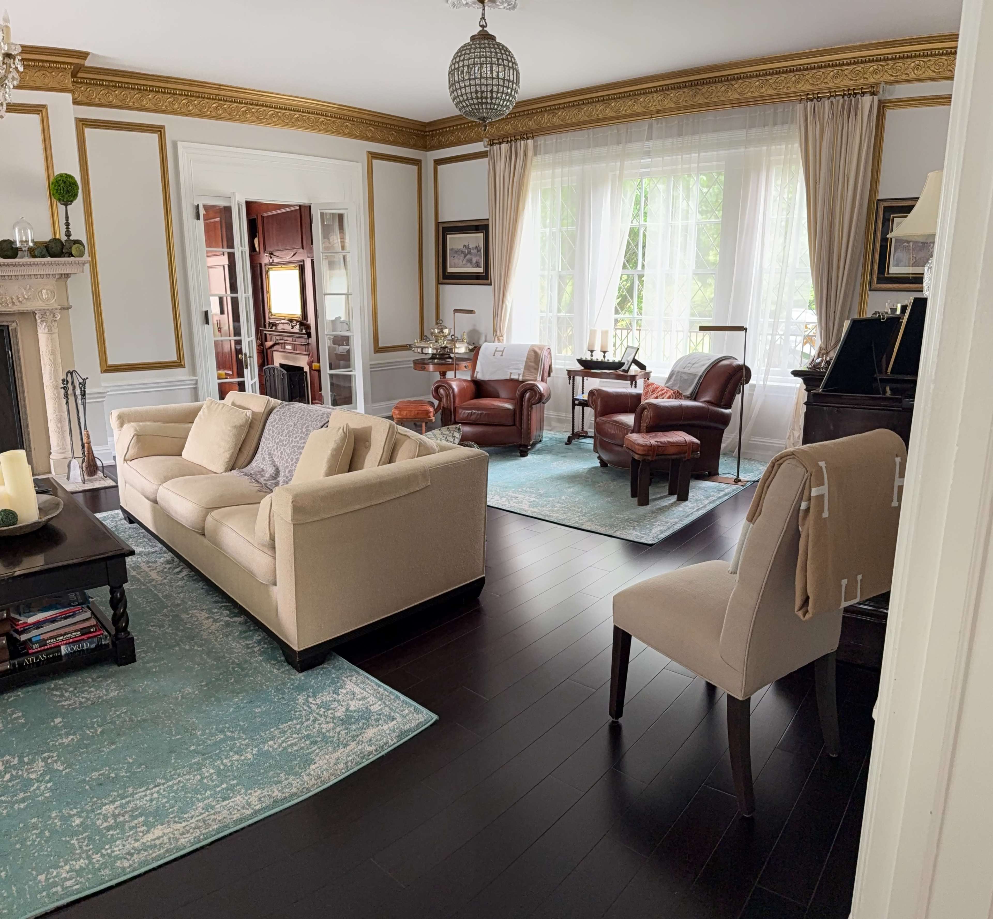The living room features a beige sofa, two leather armchairs, and a neutral-toned chair, all arranged around blue area rugs on dark hardwood flooring, with large windows allowing natural light to enter.