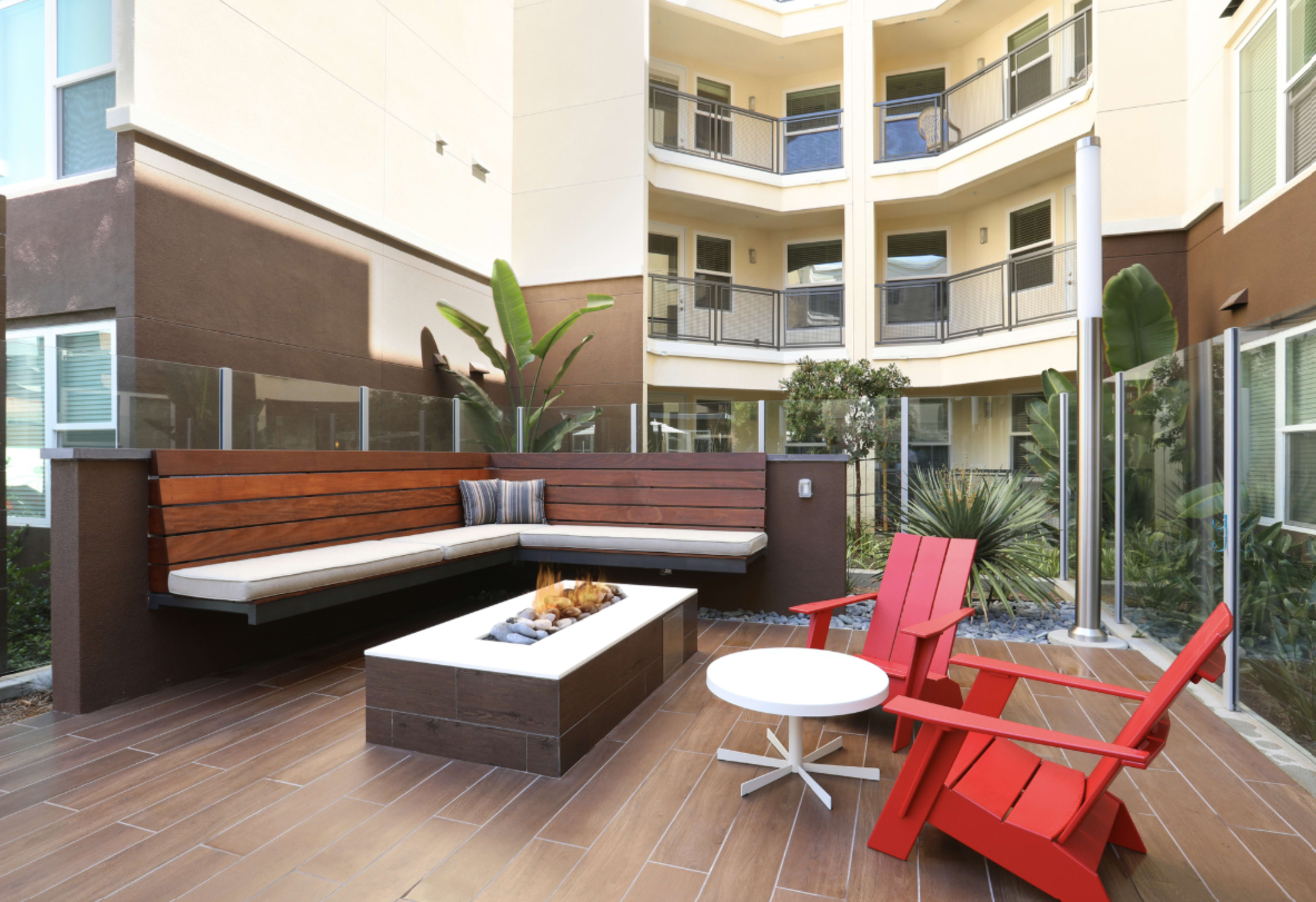The image shows a modern outdoor patio area featuring a fire pit with seating made of wooden benches and two red adirondack chairs.