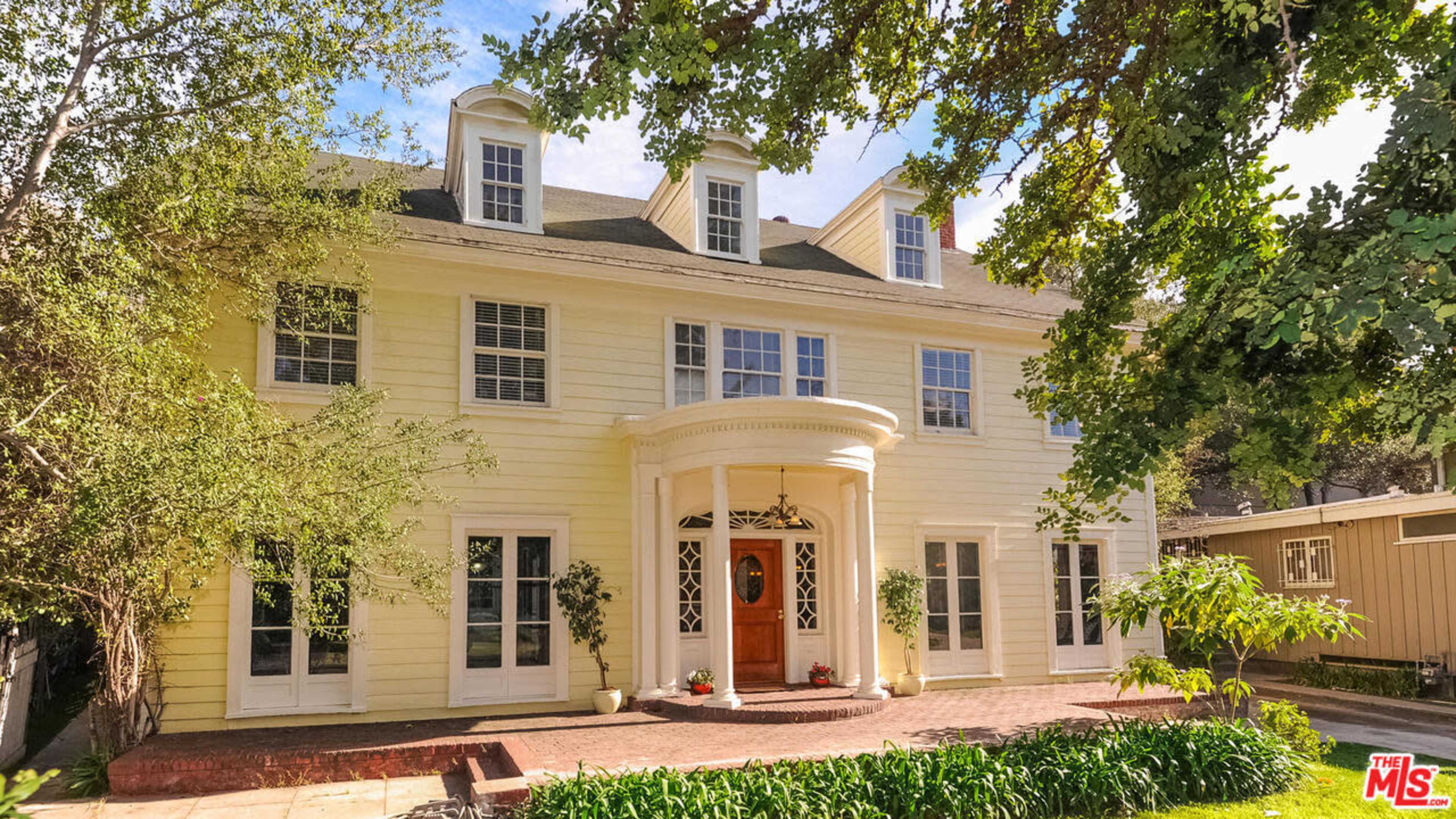 A large, two-story yellow house features a circular entryway and multiple windows surrounded by greenery.