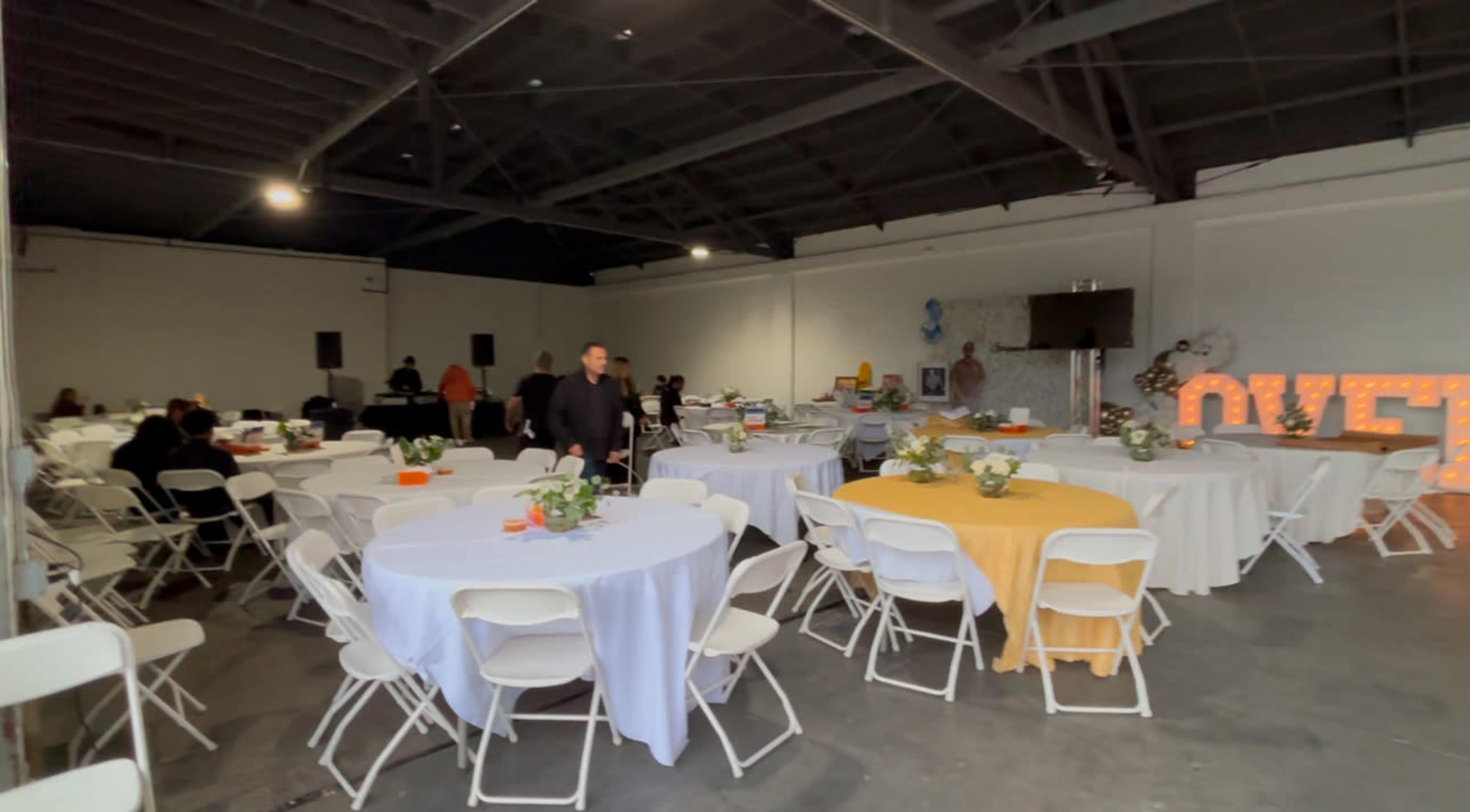 The image shows a spacious indoor event venue with several round tables set up, some covered with white tablecloths and others with yellow, and various guests mingling throughout the room.