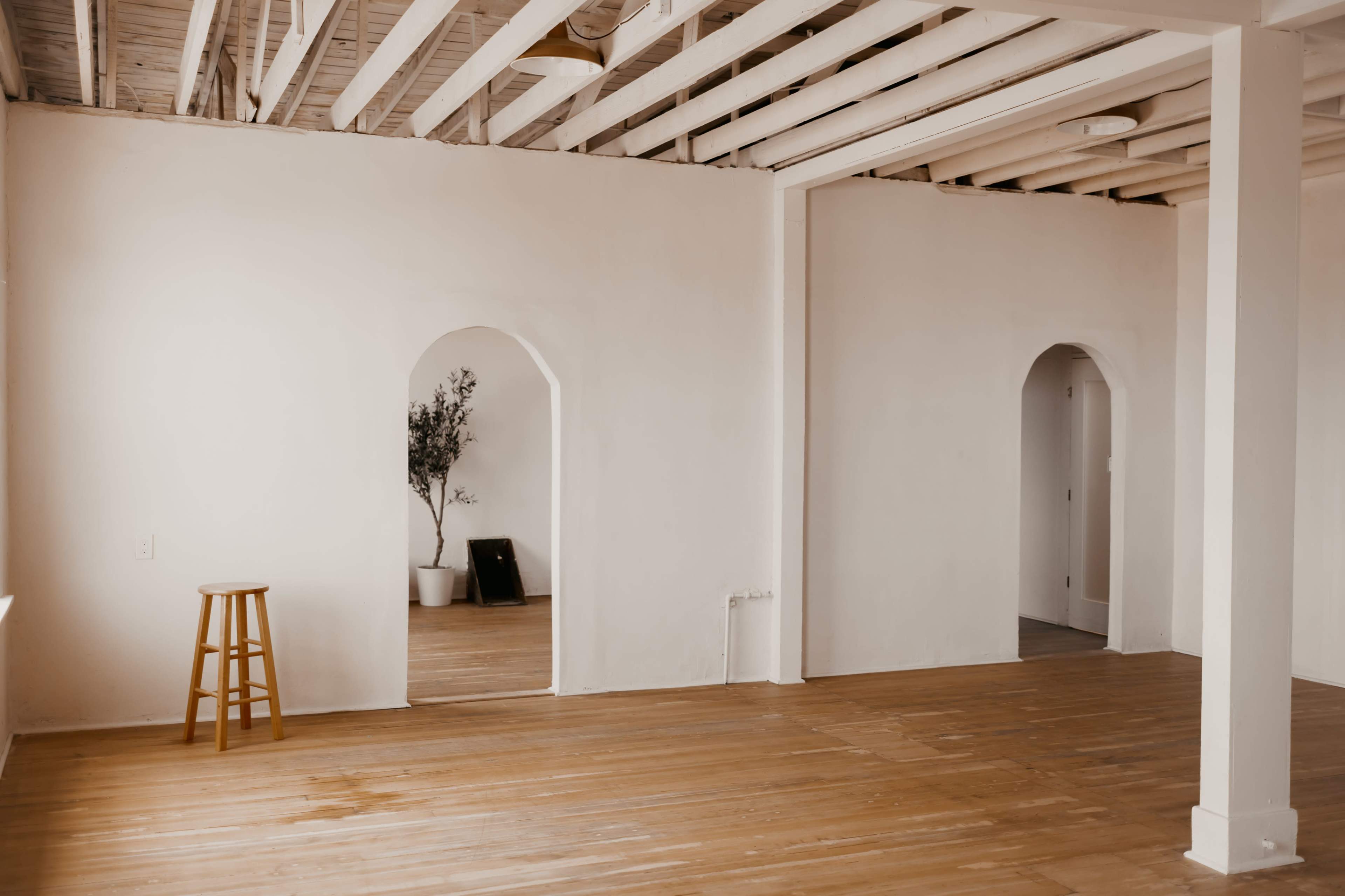 A minimalist interior space with wooden flooring and white walls, featuring archways leading to two adjoining rooms and a stool positioned near one of the arches.