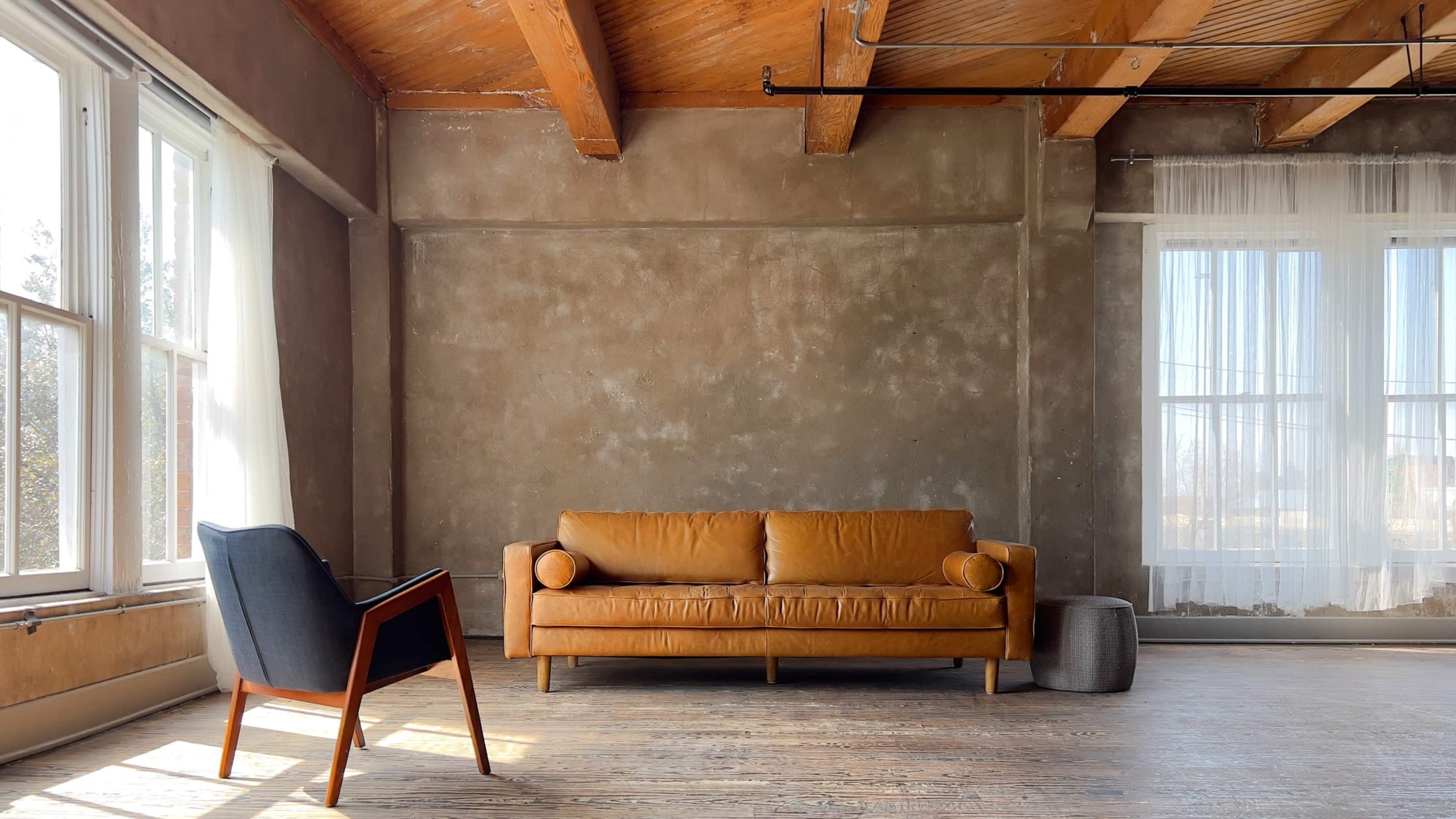 The room features a leather sofa, a modern chair, and large windows allowing natural light to illuminate the wooden floor and textured walls.