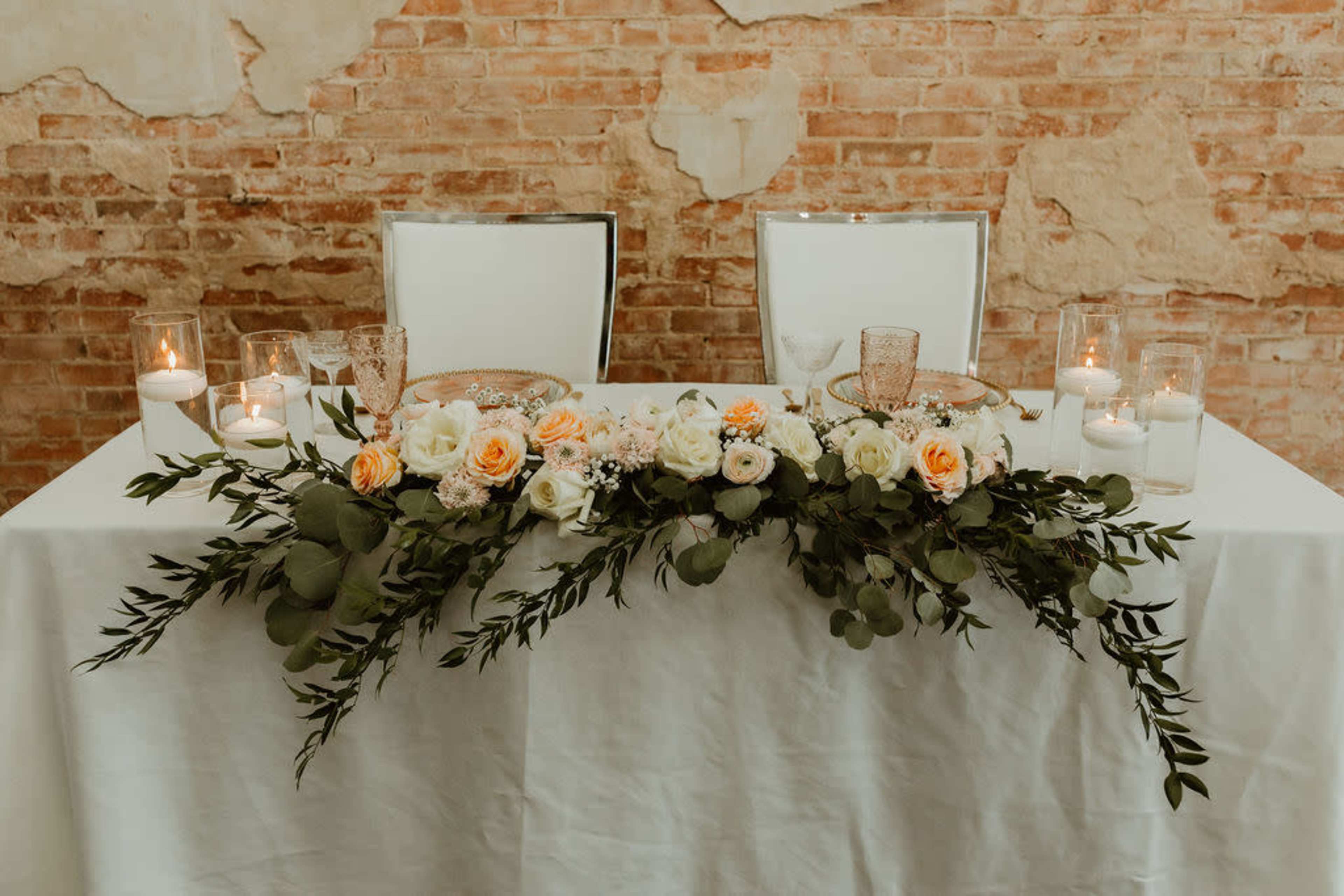 A table is decorated with a floral arrangement and candles against a rustic brick wall.