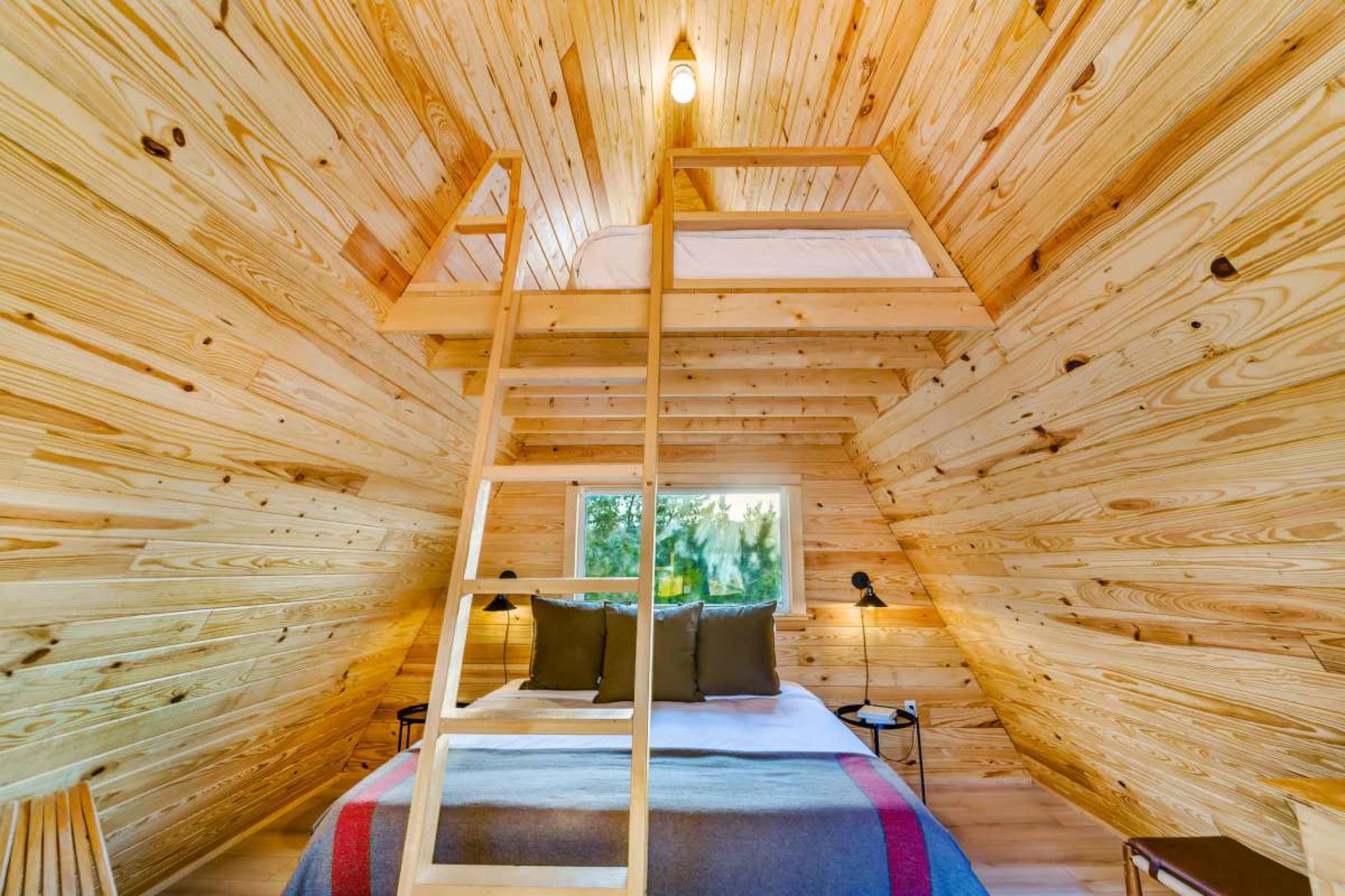 The image shows a cozy wooden interior of a small cabin with a lofted bed and a large window.