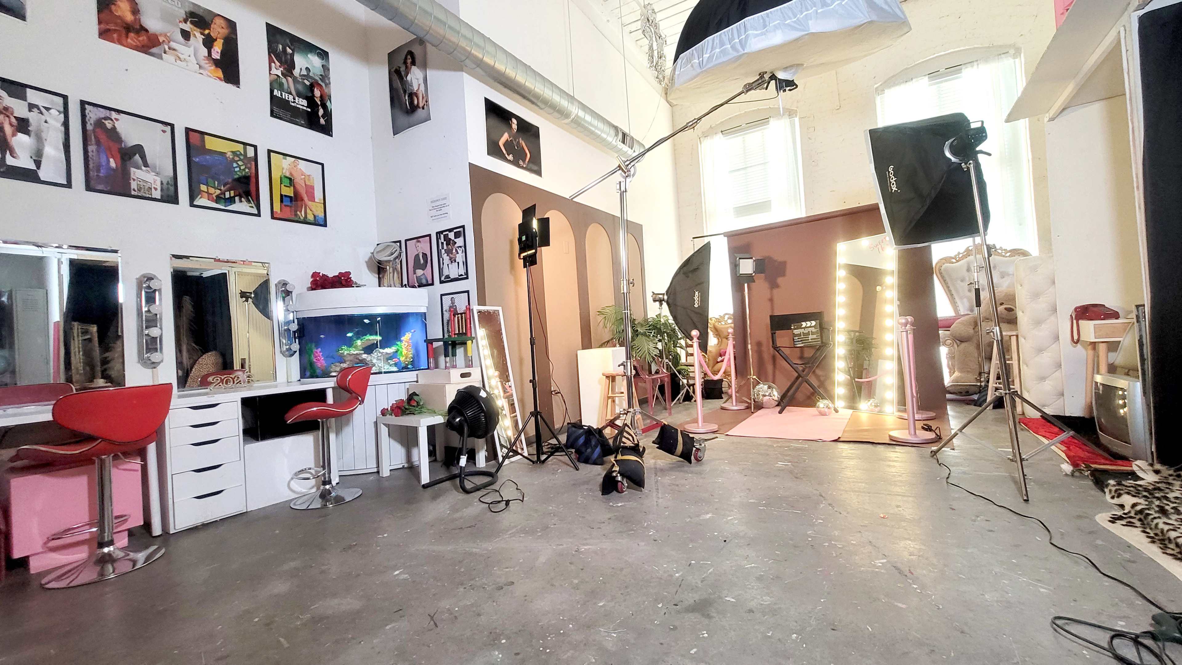The image shows a photography studio featuring a makeup area with mirrors and chairs, a backdrop with lights, and various equipment scattered on the floor.