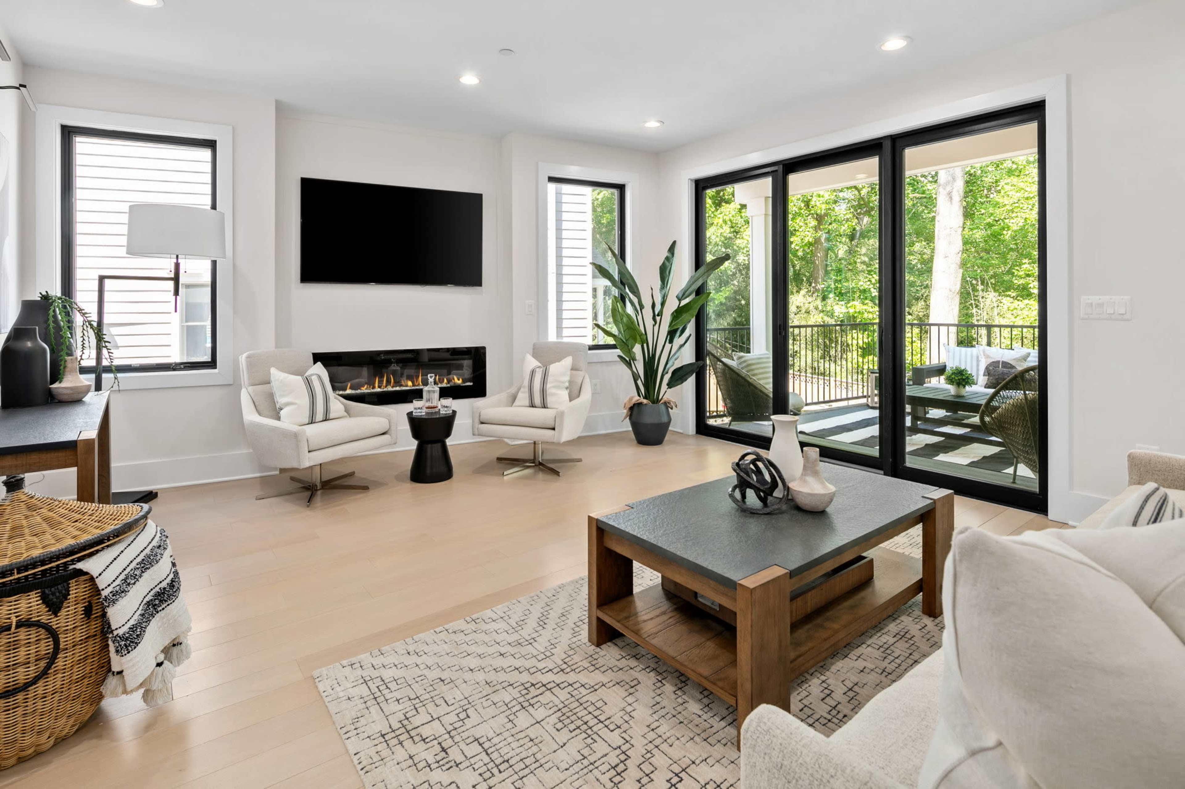 A modern living room features two armchairs, a coffee table, a fireplace, and large windows overlooking a green outdoor space.