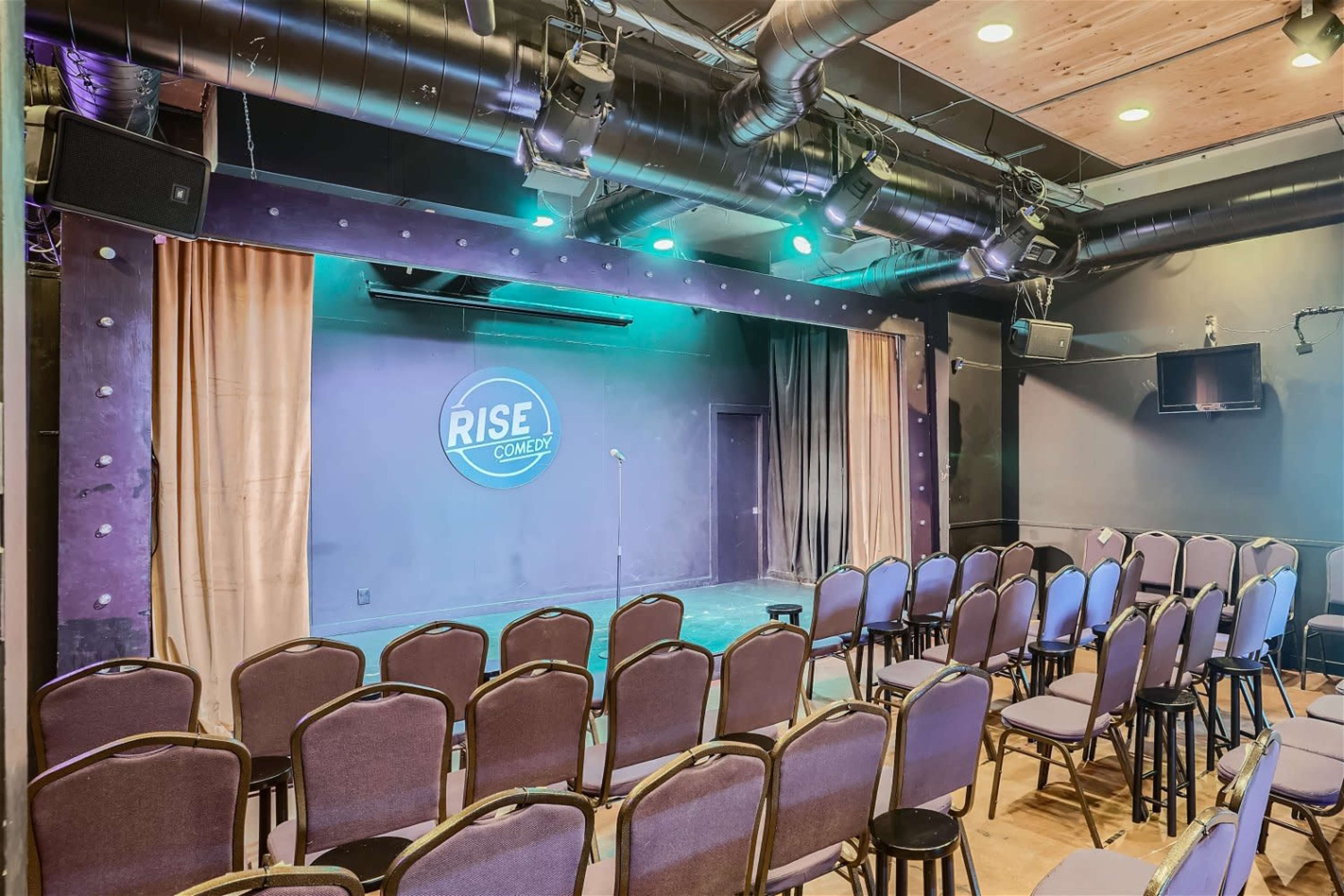 The image shows a comedy stage with a microphone and a backdrop, surrounded by rows of empty chairs.
