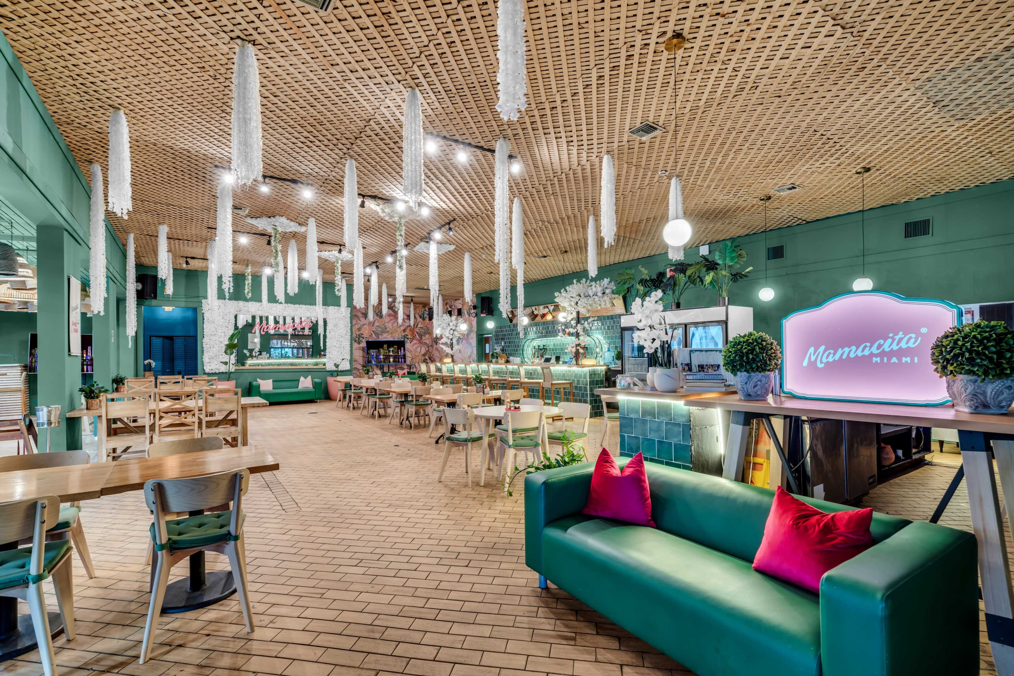 The image shows a spacious restaurant interior with green walls, wooden tables, and decorative hanging elements, featuring a bar area and a vibrant sign labeled "Mamacita Miami."
