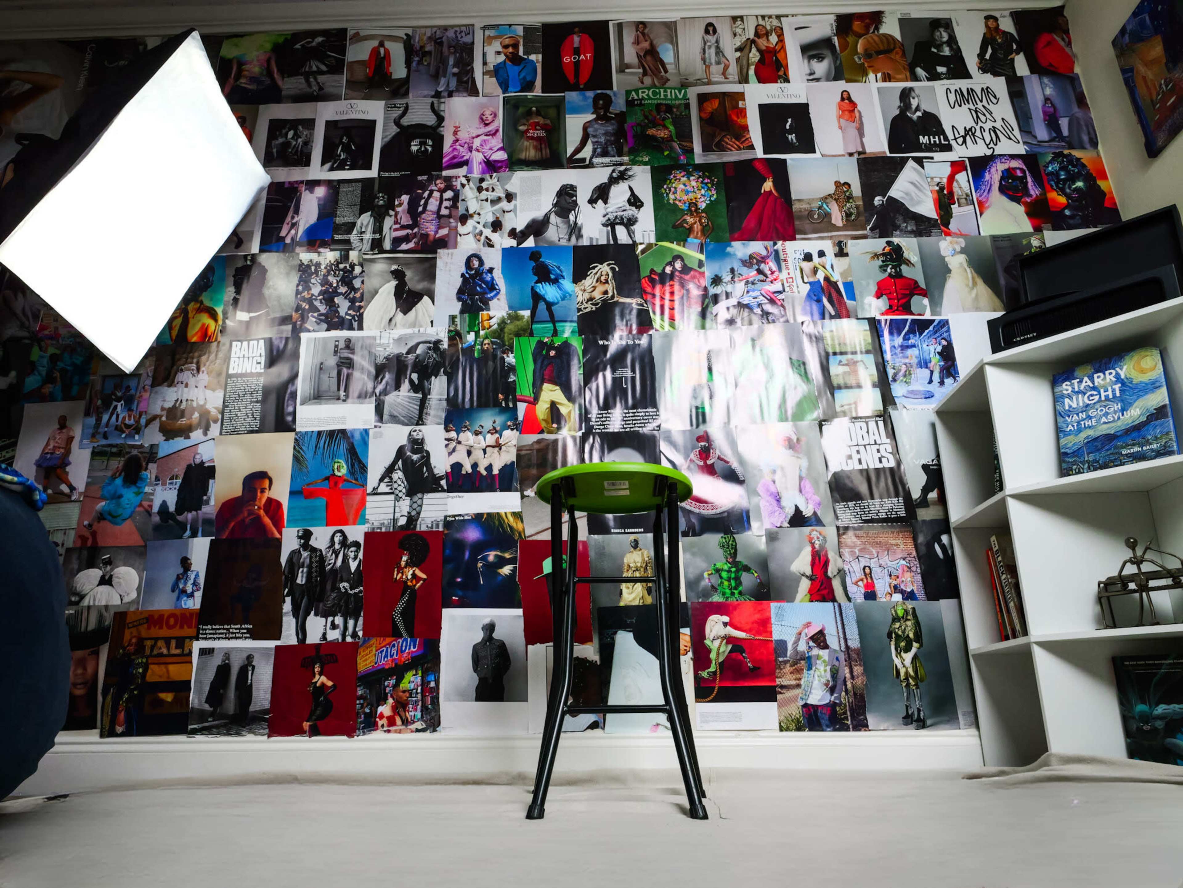The scene features a wall covered with a diverse array of photographs, a green stool positioned in front, and a light source positioned to the side.