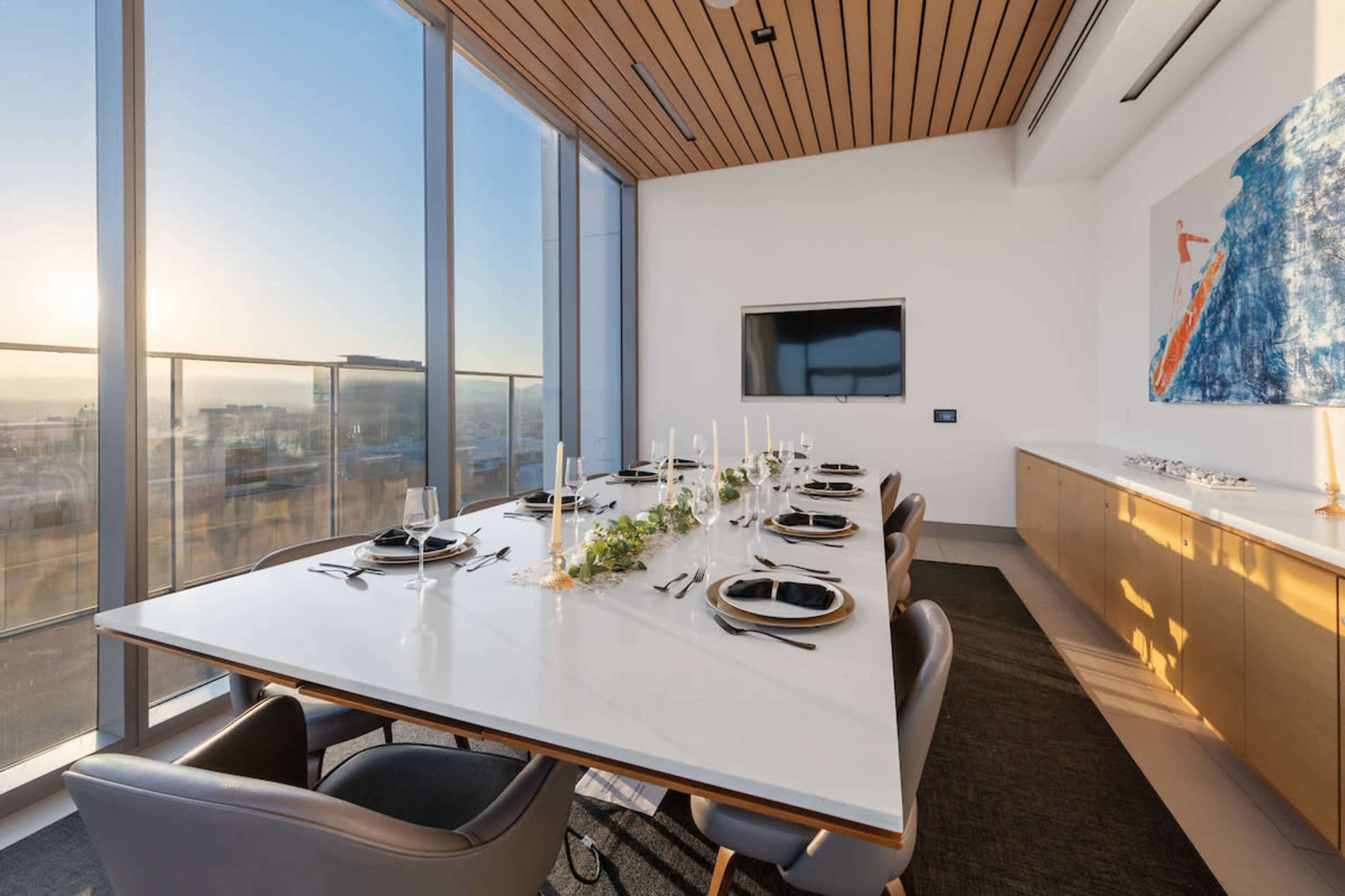 A modern dining room features a large white table set for a meal, surrounded by chairs, with floor-to-ceiling windows that provide a view of the sunset.