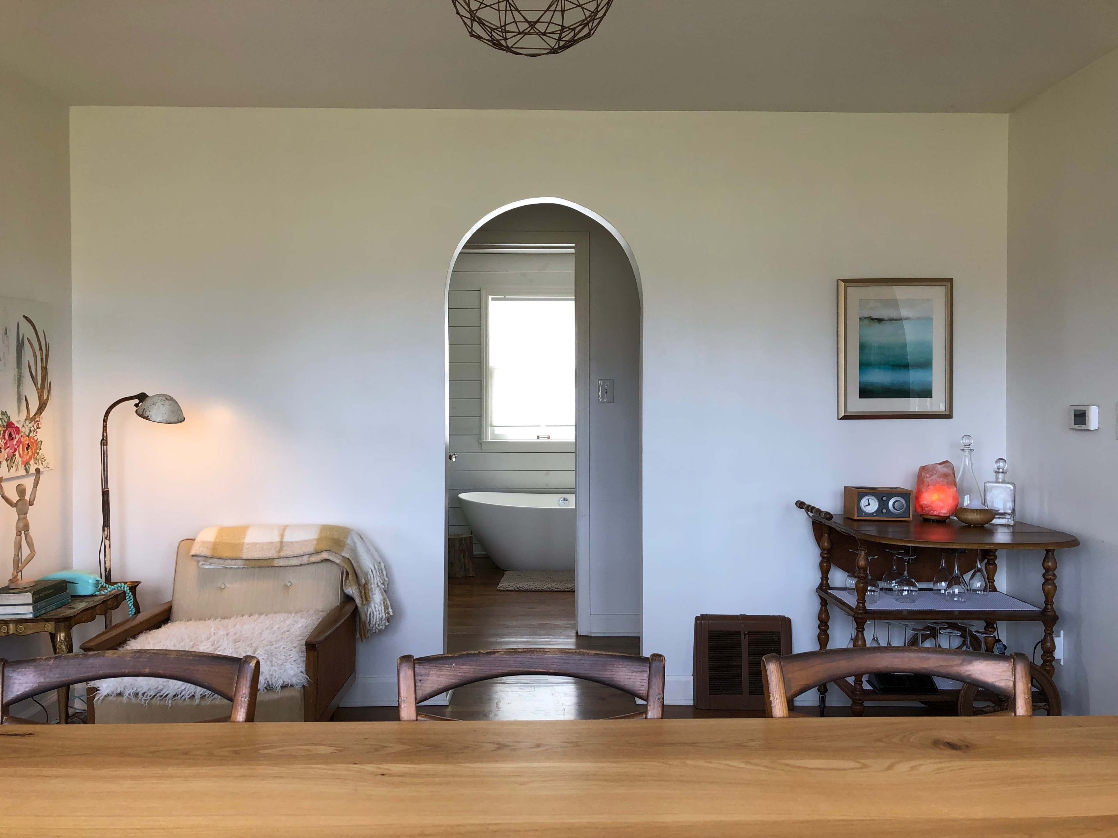 A cozy room features a wooden dining table, an archway leading to a bathroom with a freestanding tub, and a side table displaying decorative items.