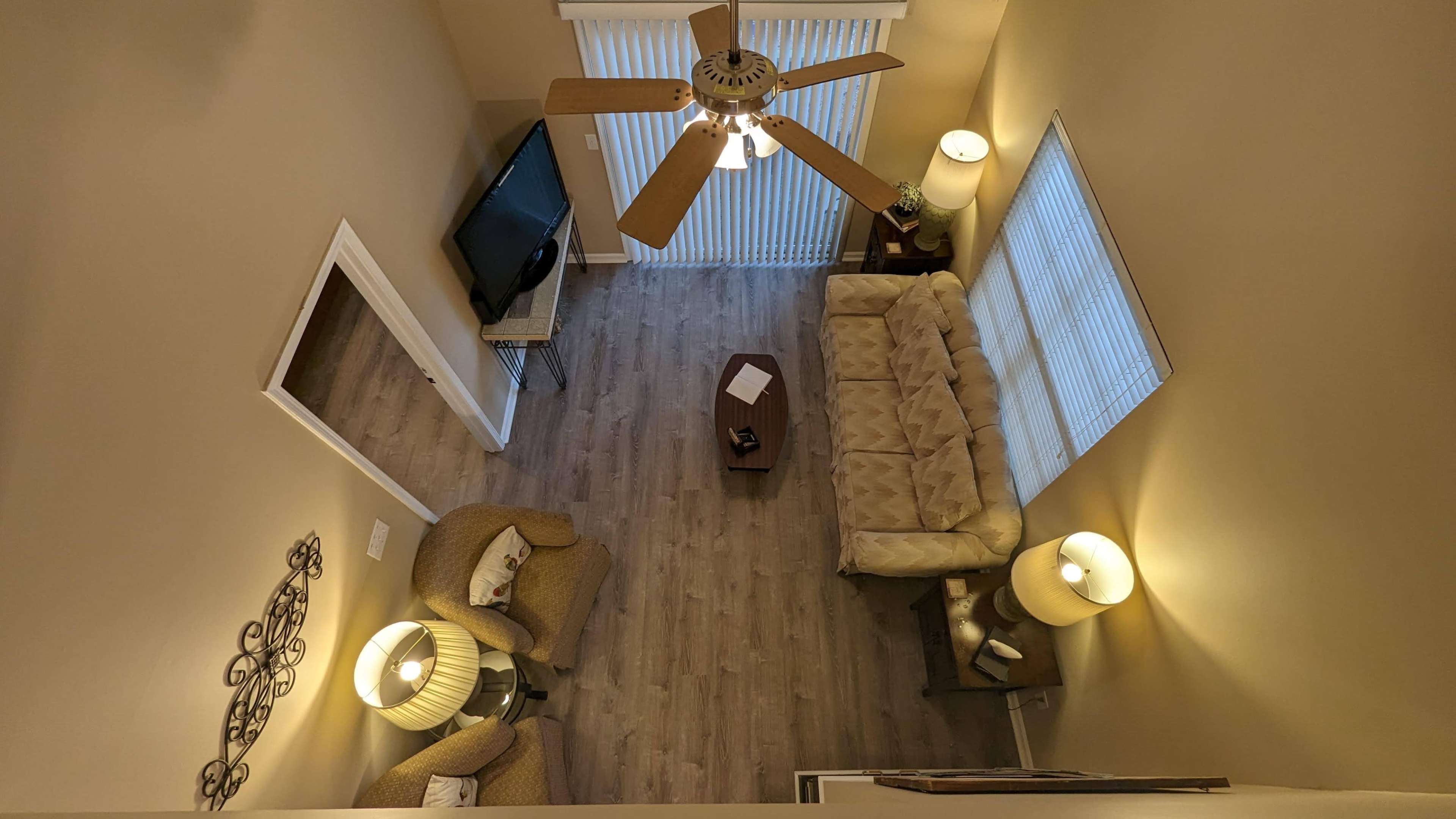A living room with a beige couch, two lamps, a television, and vertical blinds, viewed from above.