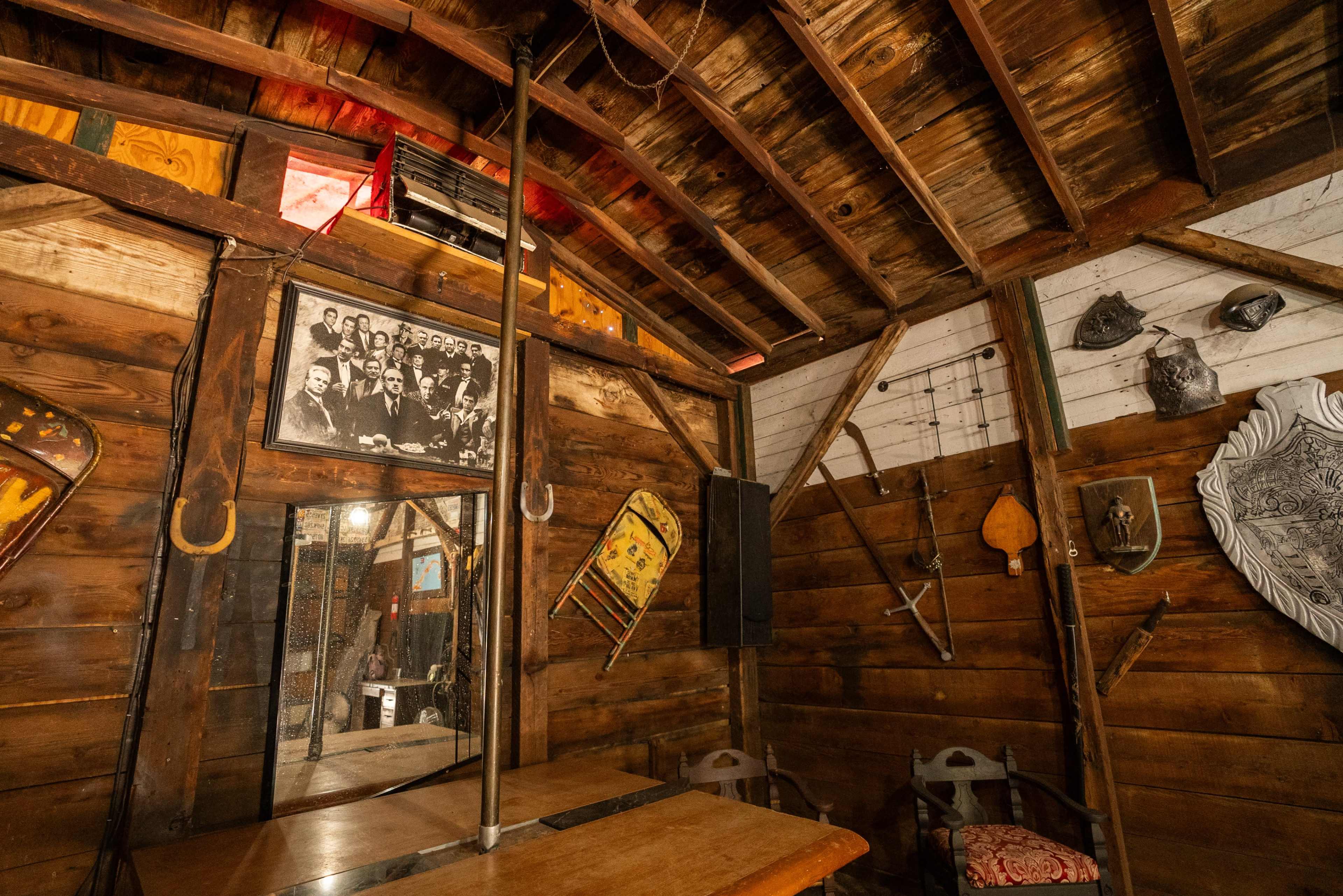 The interior of a rustic wooden structure features a wooden table, various tools and artifacts on the walls, and a mirror reflecting the surroundings.