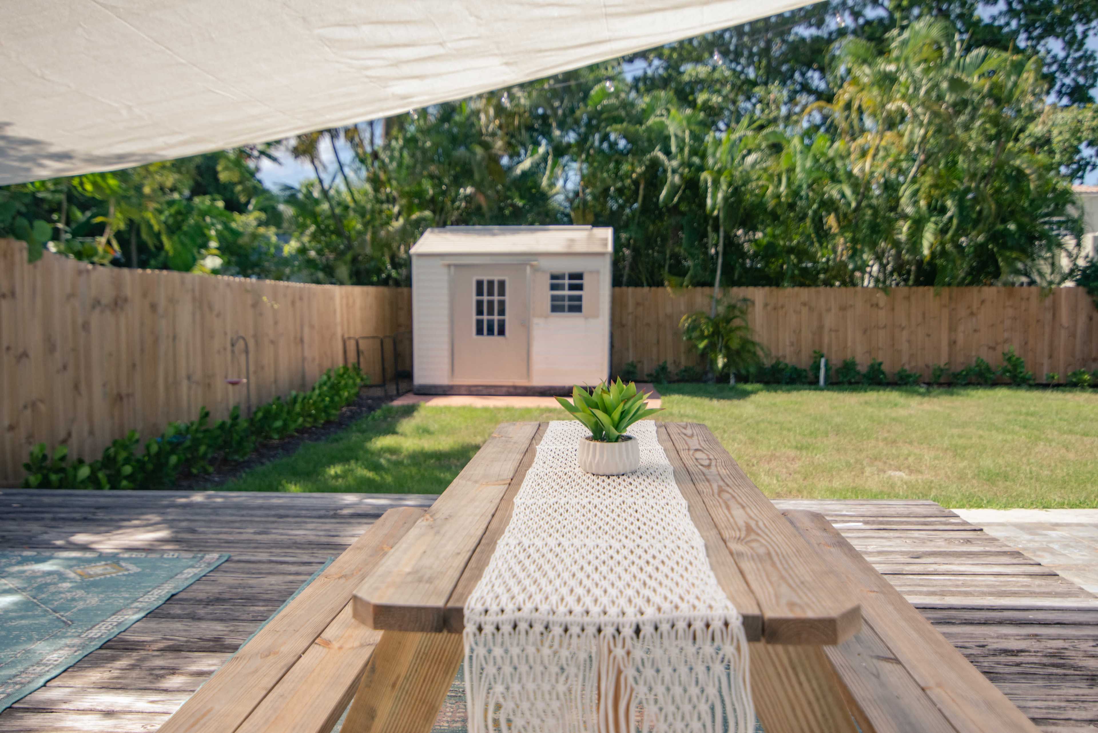 A wooden picnic table with a decorative runner is set in a backyard overlooking a small shed surrounded by greenery and a wooden fence.
