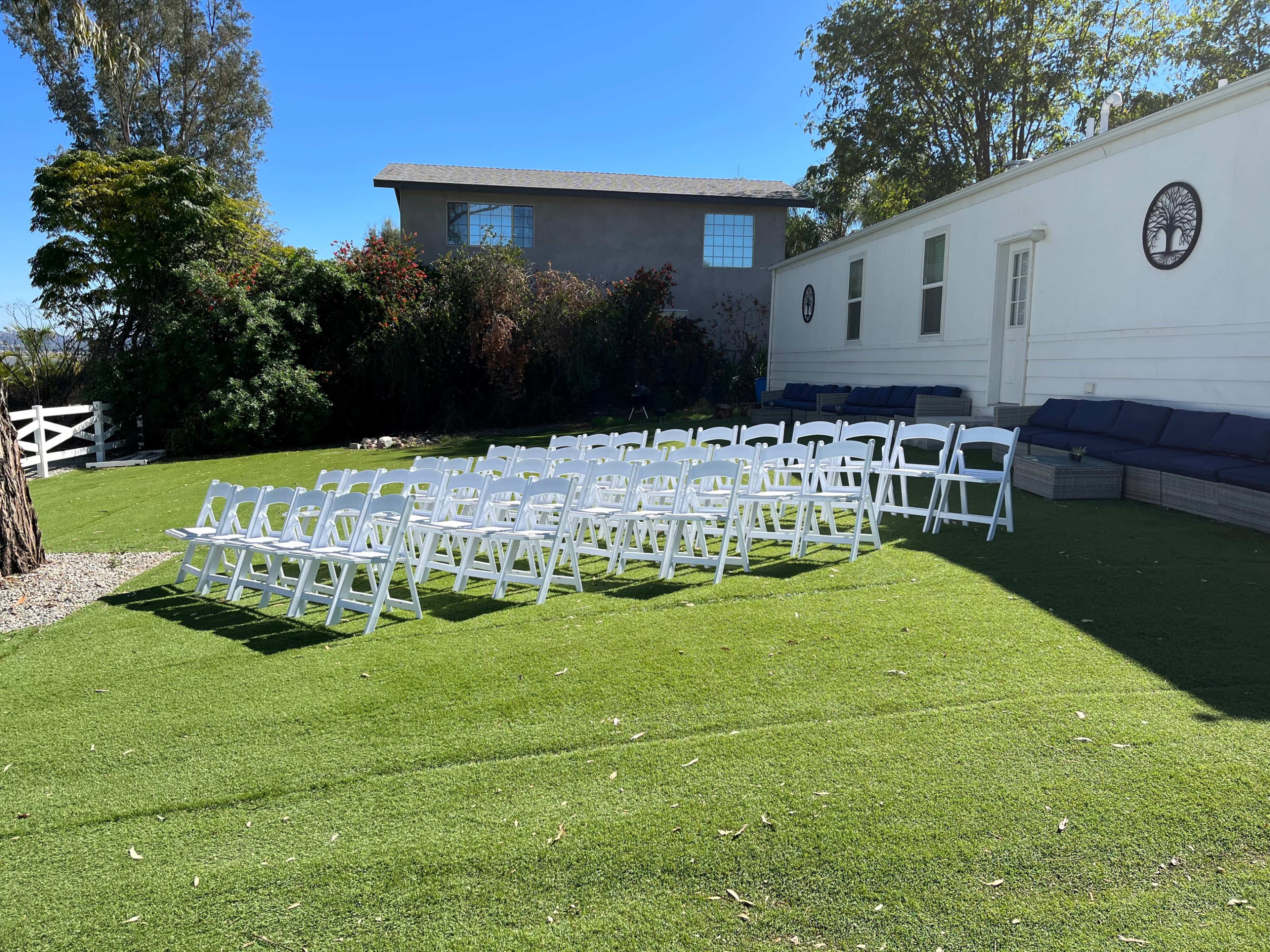 A row of white folding chairs is set up on a green lawn beside a house.