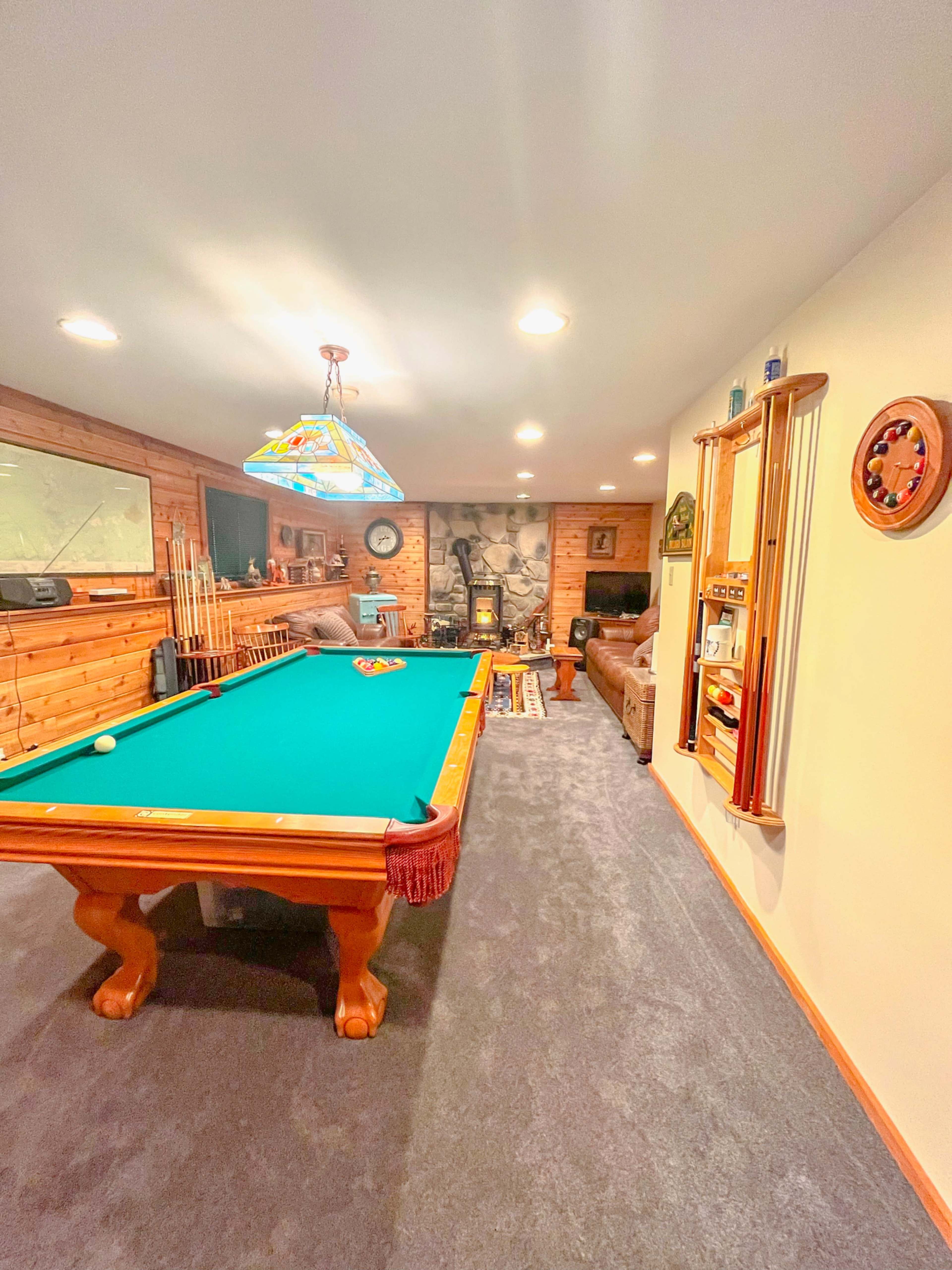 The image shows a well-lit game room featuring a green pool table, a fireplace, and various recreational equipment on the walls.