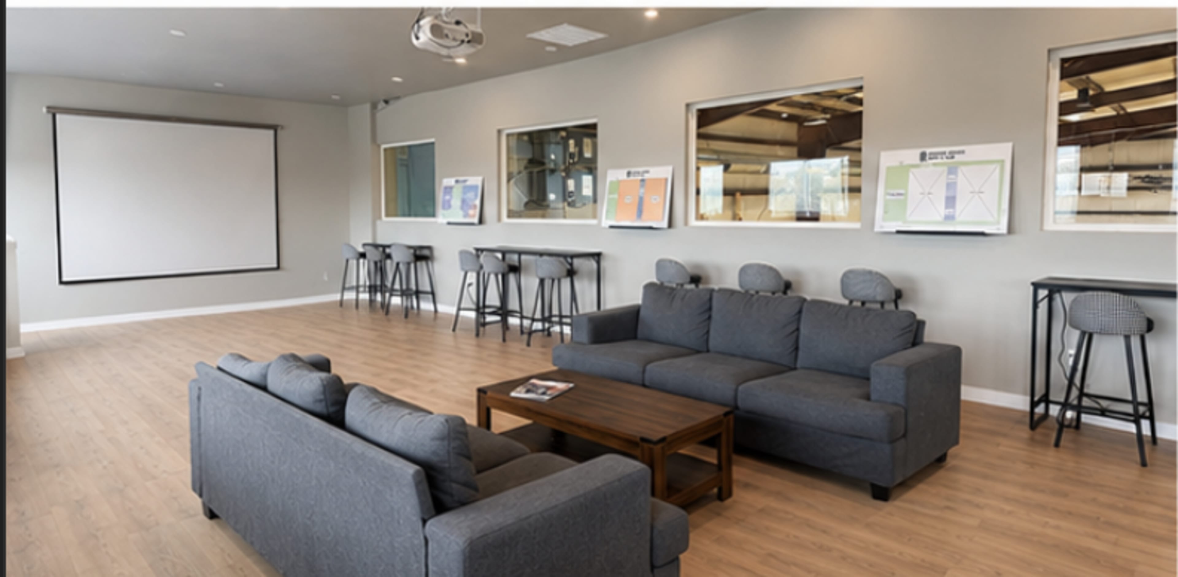 The image shows a modern lounge area with gray sofas, a coffee table, and high bar stools against a wall with windows and a projection screen.