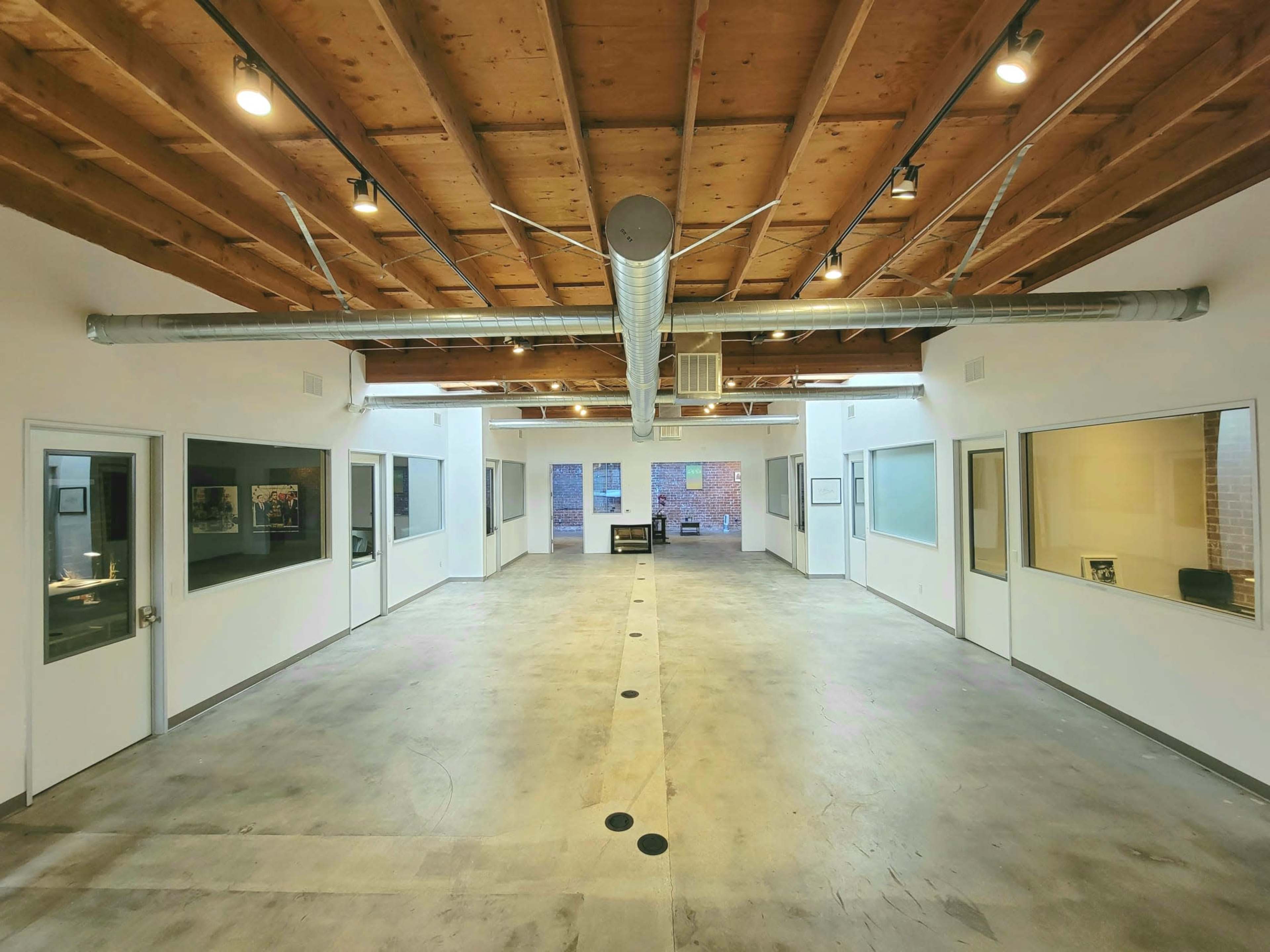 The image shows a spacious interior of an office with exposed wooden beams, concrete flooring, and several glass-walled rooms on either side.