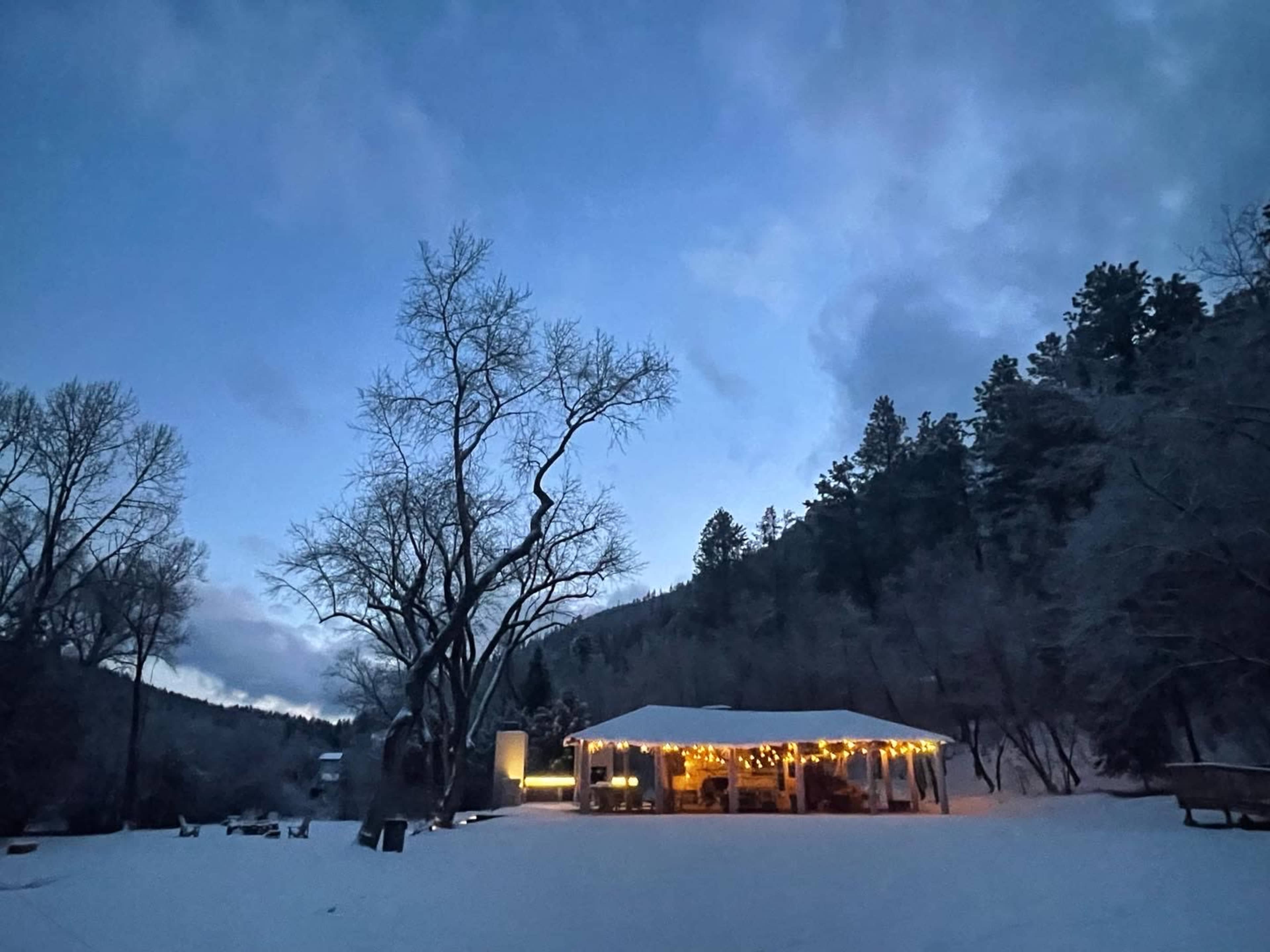 A cabin with string lights sits in a snowy landscape surrounded by trees under a twilight sky.