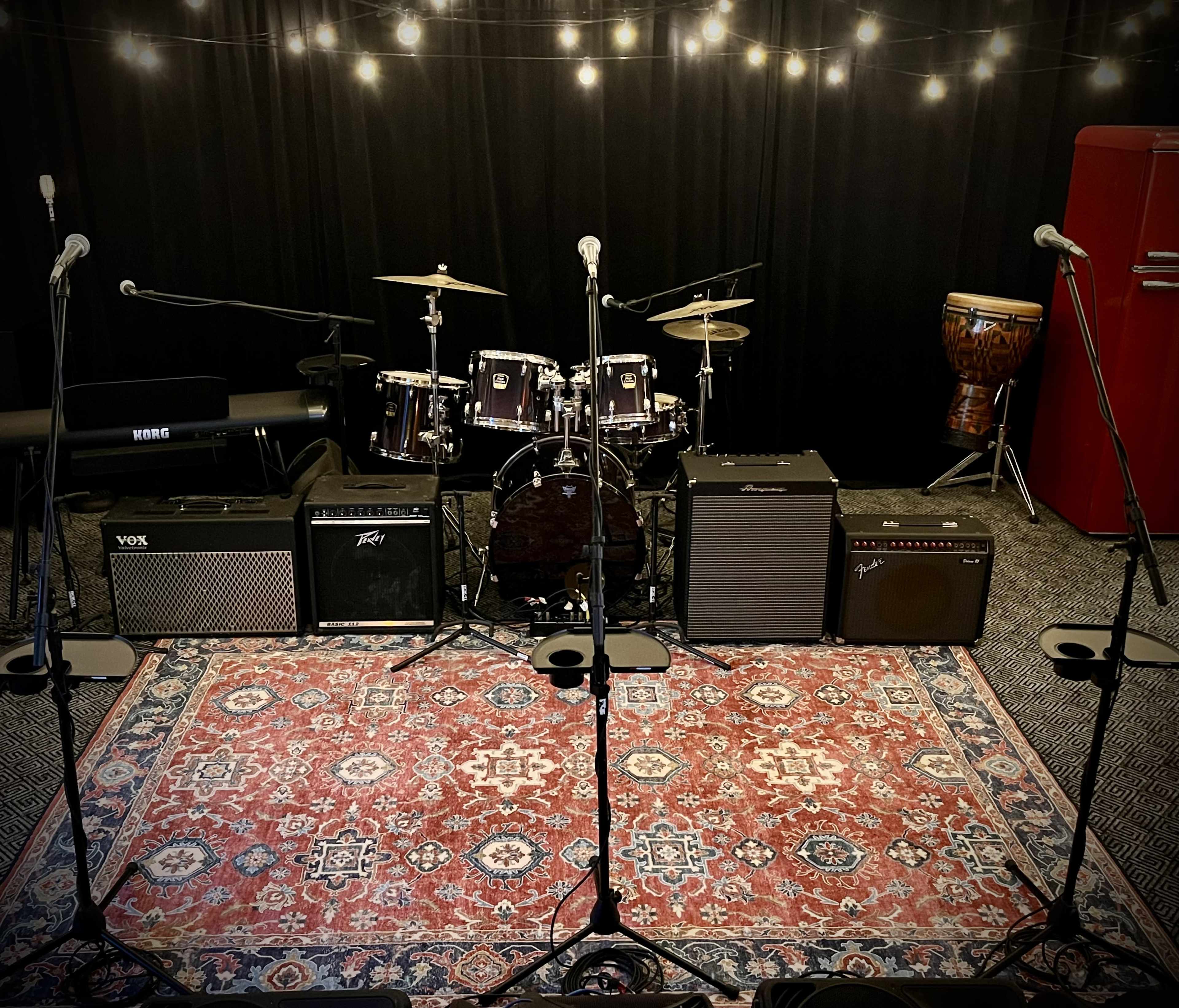 The image shows a stage setup with musical instruments, including a drum kit, amplifiers, microphones, and a keyboard, arranged in front of a black backdrop adorned with string lights.